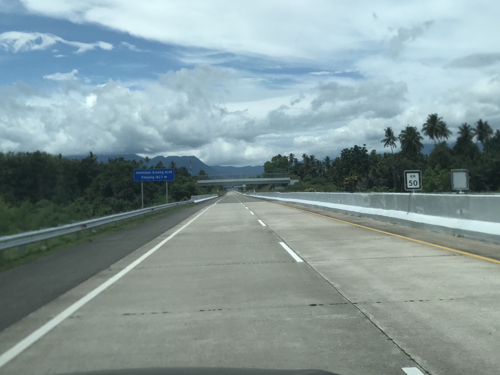 Captured from inside a vehicle, the image presents a view of a straight, gray concrete highway in Seuot Tunong, Indonesia. The road, divided by white lane markings and a yellow shoulder line, stretches toward a distant overpass. A blue sign on the left indicates "Jembatan Krueng Aceh Panjang 162.7 M," denoting the name and length of a bridge, while a "KM 50" sign appears on the right, indicating distance. Green foliage and trees line both sides of the road, with coconut palms visible on the right. A mountain range is faintly discernible in the background under a sky filled with white and gray clouds. The vehicle's dashboard is partially visible at the bottom of the frame. The road appears to be clear of traffic.