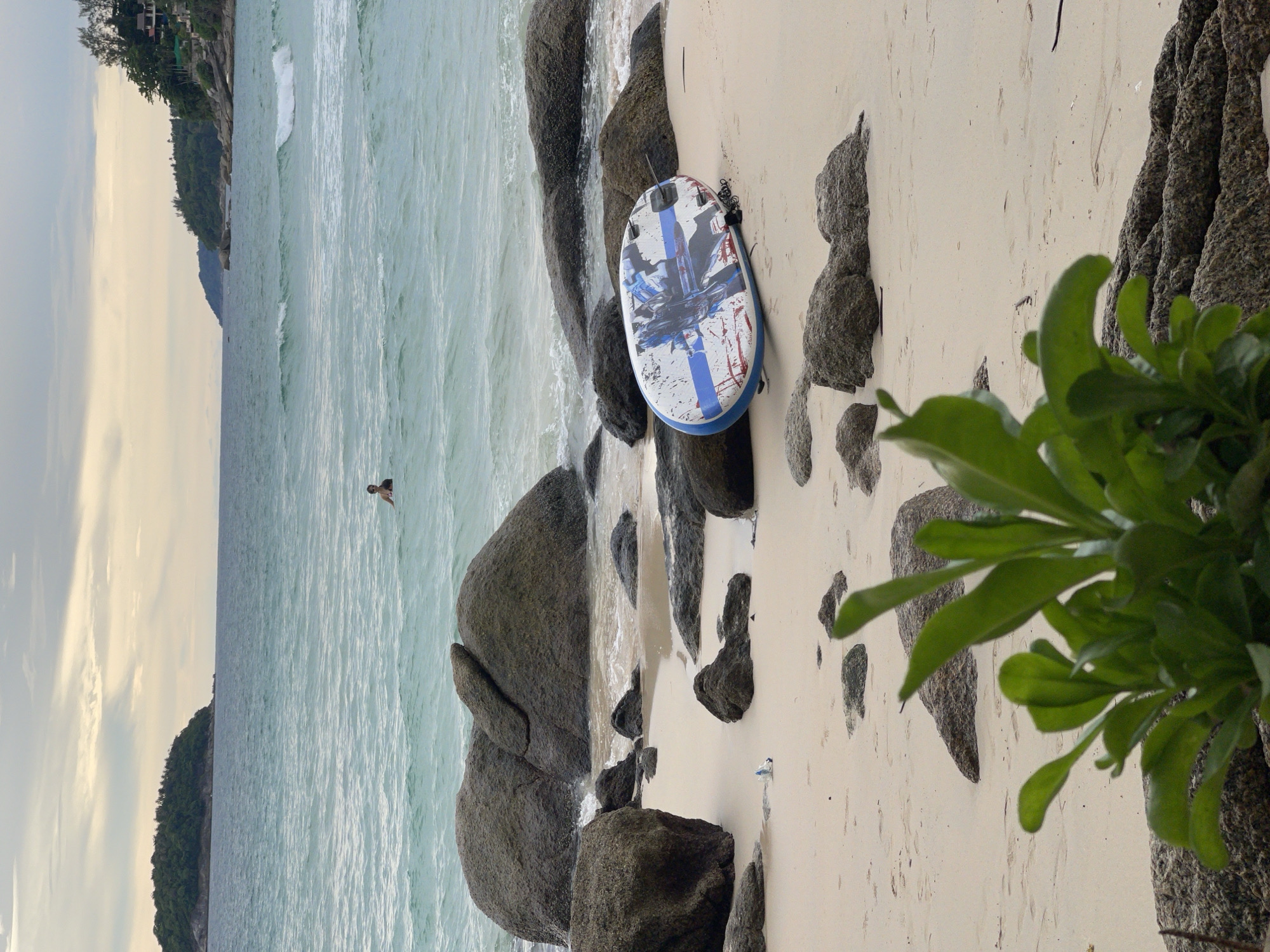 The image depicts a serene beach scene in Karon, Thailand, with a person swimming in the turquoise waters. A weathered paddleboard lies on the sandy shore, its surface adorned with a blue and white graphic design, hinting at past recreational activities. Large, dark rocks dot the beach, some partially submerged in the gentle waves, while a patch of lush green foliage in the foreground adds a touch of natural beauty. The sky overhead is overcast, casting a soft, diffused light on the landscape. The overall atmosphere is one of tranquility and natural splendor.