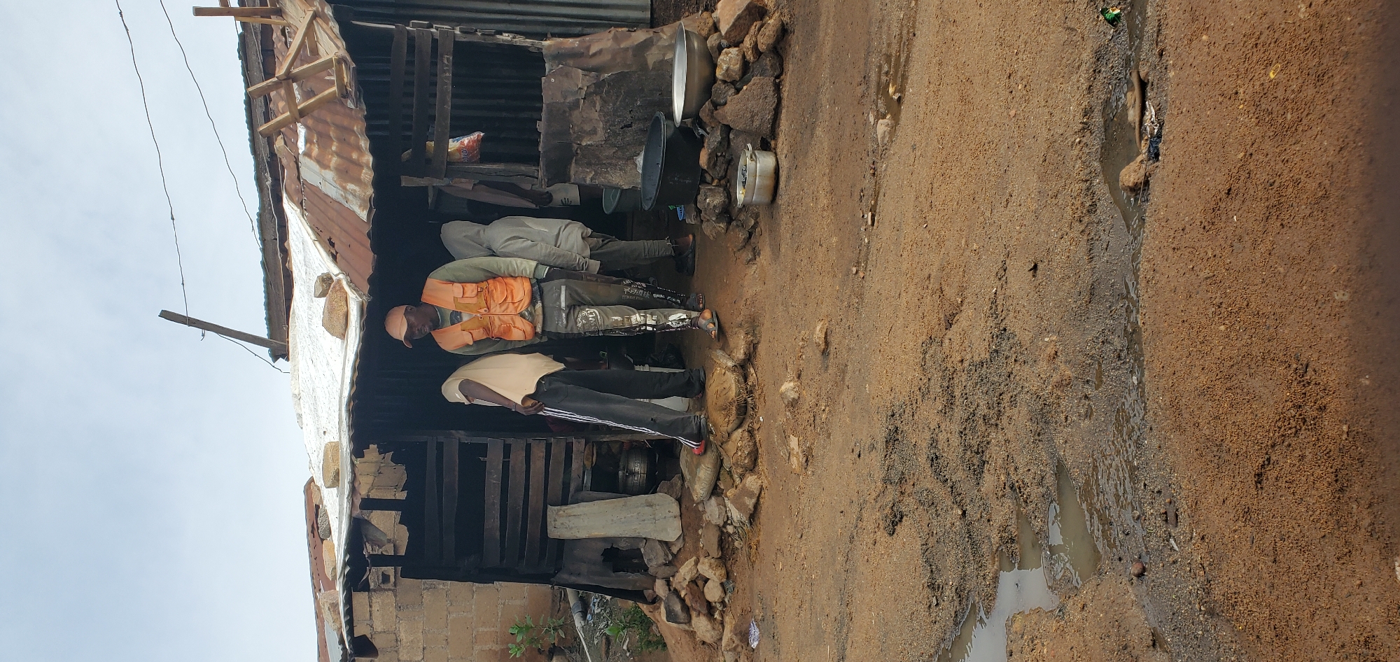 Four individuals are standing in front of a makeshift structure with a corrugated metal roof. One man in the foreground wears an orange vest over a green long-sleeved shirt and patterned pants. To his left, a person in a light-colored shirt and dark pants sweeps the ground with a broom. Behind them, two other individuals are visible, one wearing a grey hooded sweatshirt. The structure appears to be made of wood and corrugated metal, with visible repairs using rocks and other materials. Several metal pots and basins are placed outside the structure. The ground in front of the building is dirt and gravel, with puddles of water and tire tracks visible. The scene suggests an outdoor market stall or a simple dwelling in a rural or developing urban environment.
