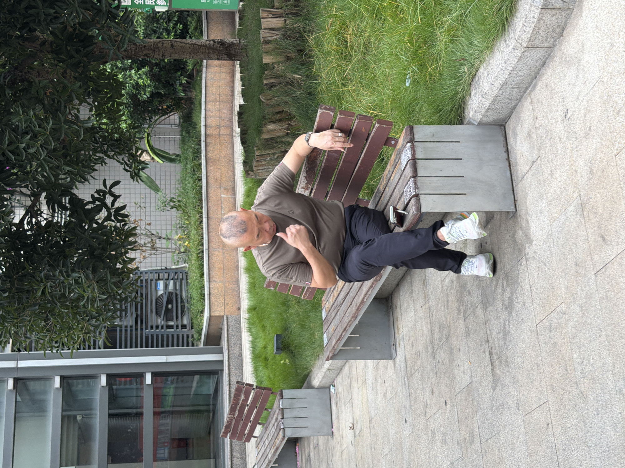 A middle-aged East Asian man with a shaved head is seated on a park bench made of weathered wood and concrete. He is wearing a dark olive t-shirt, black pants, and white and green athletic shoes. His right arm is resting on the back of the bench, and he appears to be looking down at his phone, which is on the bench beside him. He has a ring on his left ring finger and a watch on his left wrist. The bench is positioned on a paved walkway in what appears to be a public park or outdoor area in Yuzhong District, China. To the left of the man is another empty bench. Behind the benches, there is a manicured lawn and several trees, including a larger tree with a textured bark. In the background, a modern building with glass windows and an air conditioning unit is visible. The overall scene suggests a casual moment of rest in an urban outdoor environment.