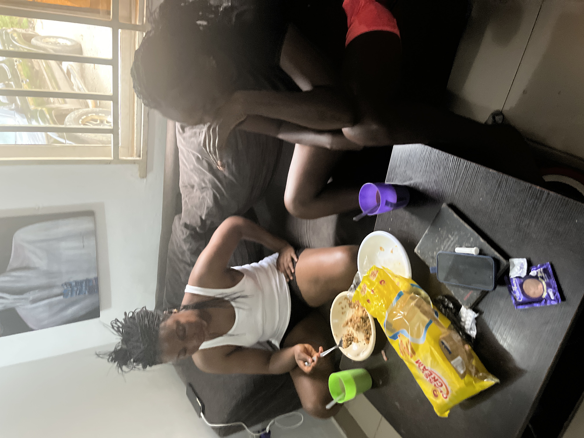 Two women are seated on a dark brown couch in an indoor setting, likely a living room in Abuja, Nigeria. The woman on the left, wearing a white tank top and dark shorts, is actively eating from a bowl on a dark wooden coffee table in front of her. She holds a spoon in her right hand and appears to be mid-meal. To her left, on the table, are a green plastic cup, an open package of biscuits labeled "CREAM," and a plate of food. A dark-colored smartphone lies on a book to the right of the biscuit package. Further to the right on the table is a purple cup.
The woman on the right, dressed in a dark top and red shorts, is partially visible, her face obscured as she rests her head in her hands, facing away from the camera. Her body language suggests contemplation or distress. The background features a dark brown couch and a wall adorned with a painting of a figure in white attire. A window in the background reveals a glimpse of an outdoor scene with a vehicle. The lighting is moderate, with shadows present, indicating it is likely daytime.