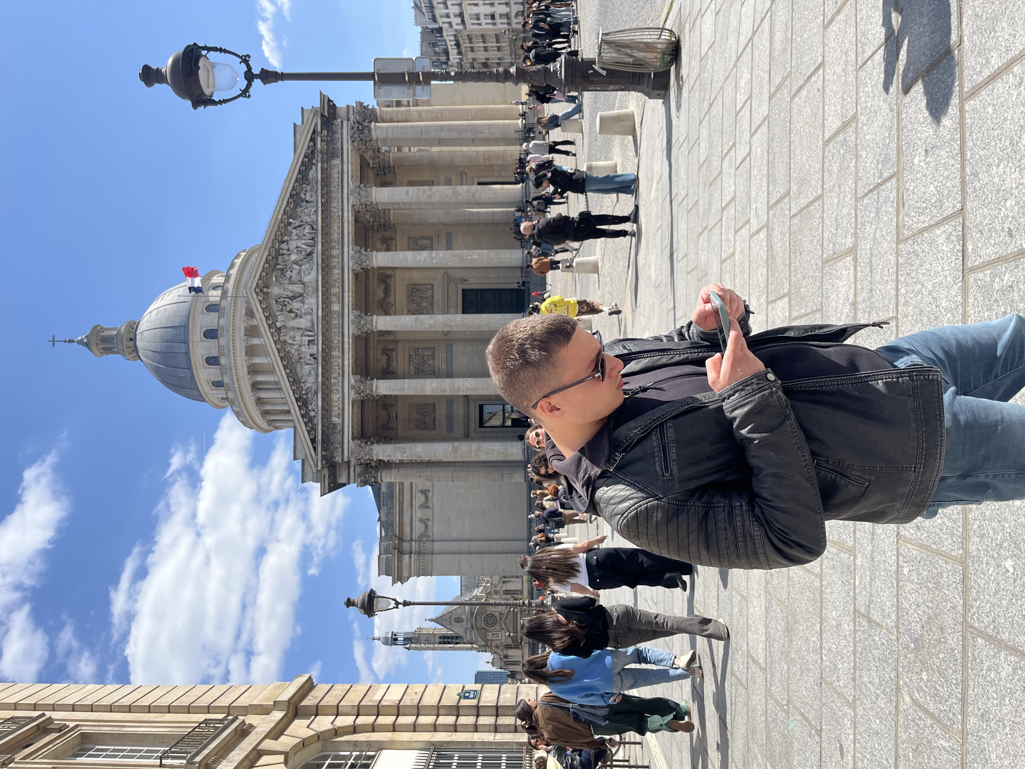 The image captures an outdoor urban scene on a bright, sunny day, likely in the mid-morning or afternoon due to the strong shadows cast. The primary subject in the foreground is a man, viewed from the side, wearing a black leather jacket, a dark shirt, and blue jeans. He is wearing sunglasses and is engrossed in looking at a light blue smartphone held in his hands.
Behind the man, a grand, neoclassical building dominates the background. This building features a large dome topped with a finial and a cross, and a prominent portico with numerous Corinthian columns supporting a triangular pediment. A French tricolor flag flies from the base of the dome. The pediment bears a visible inscription, "AUX GRANDS HOMMES LA PATRIE RECONNAISSANTE," which identifies the building as the Panthéon in Paris, France.
Numerous other people are scattered across the paved square in front of the Panthéon, some walking, others standing or congregating, appearing as pedestrians or tourists. The pavement consists of light grey square tiles. Streetlights and bollards are also visible in the public space. In the far background to the left, another building with a distinctive spire can be seen, likely the Church of Saint-Étienne-du-Mont, located near the Panthéon. The sky is clear blue with some scattered white clouds. The overall atmosphere is lively and typical of a public square in a major European city.