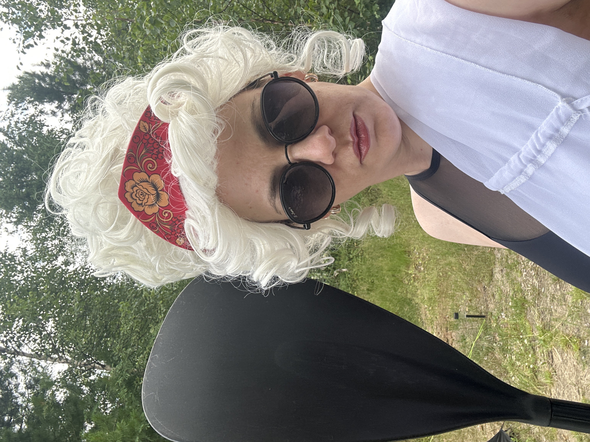 A person, possibly female, stands outdoors in what appears to be a natural setting with trees and grass. They are wearing a white, curly wig with a red and gold floral headband. Round, dark sunglasses are on their face, and their lips are painted with a glossy lipstick. A sheer white garment is draped over their right shoulder. To the left of the person, a portion of a large, black paddle is visible. The background consists of greenery and a cloudy sky. The specific location in Tolbazikha, Russia is not discernible from the image.