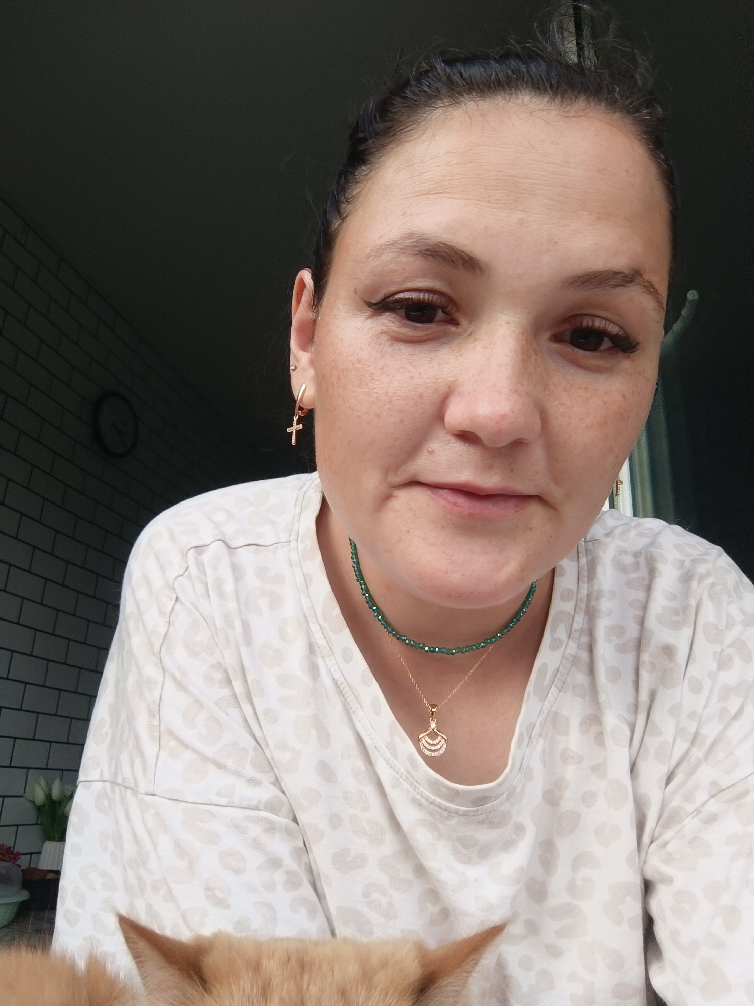 The image shows a close-up, eye-level portrait of a woman with dark hair pulled back and a light-colored, leopard-print t-shirt. She is wearing a thin green beaded necklace with a gold pendant and a gold cross earring in her left ear. The background is dark, with a hint of a tiled wall and a clock on the left side. A ginger cat's head is visible at the bottom of the frame, partially obscuring the woman's chest. The scene appears to be indoors, possibly in a residential setting in Chaikovsky, Russia.