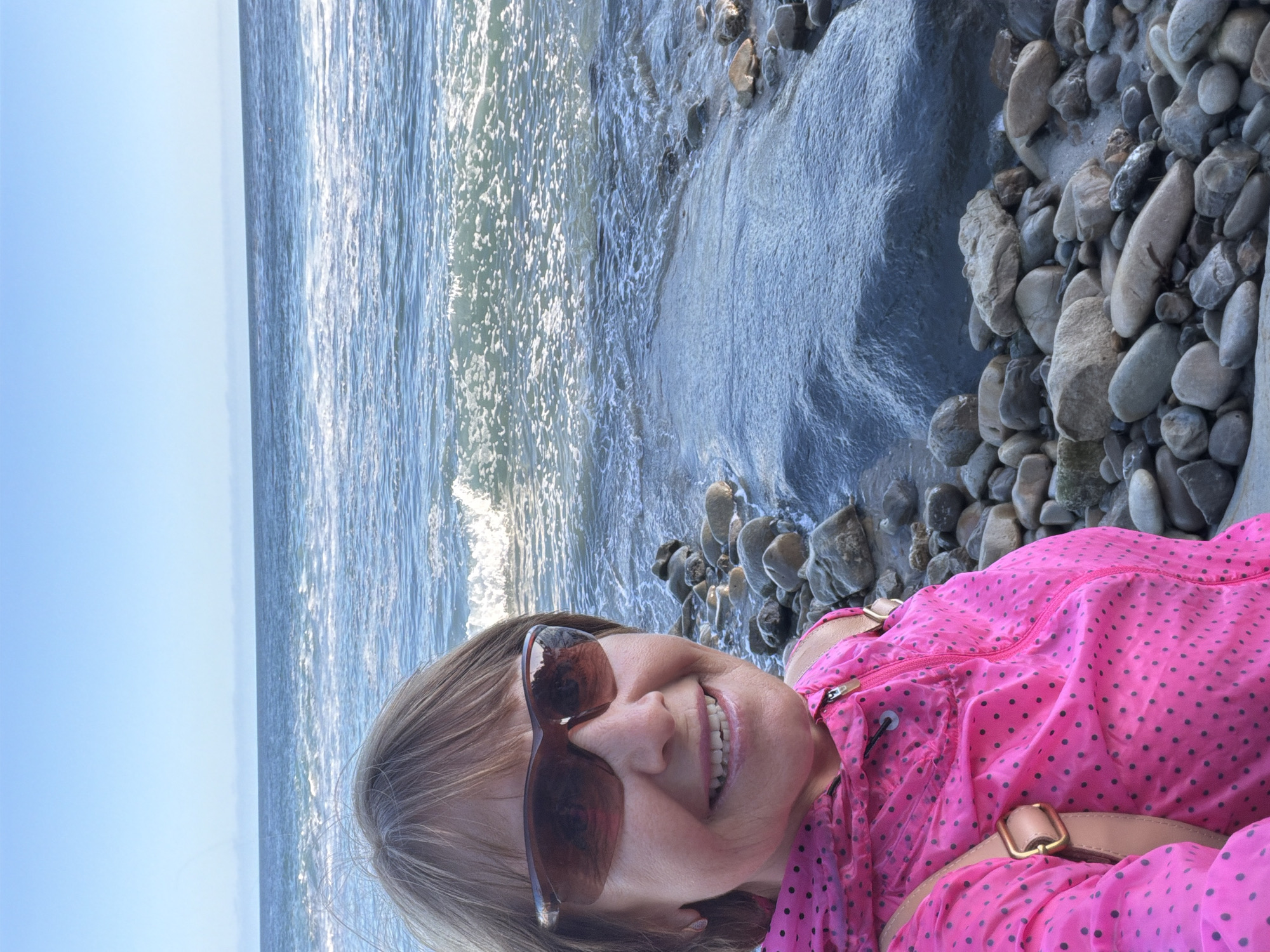 A woman with short brown hair, wearing sunglasses and a bright pink polka-dot jacket, smiles at the camera. She is standing on a rocky beach with the ocean behind her. The waves are gentle, and the sky is clear and blue. The overall impression is a sunny day at the beach. The location is confirmed as Laguna Beach, United States of America.