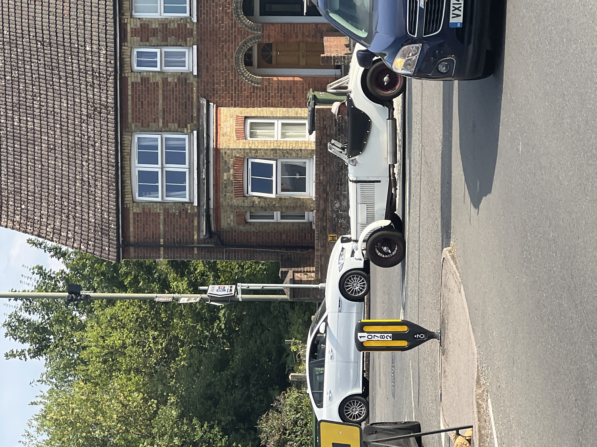 In a street scene in Guildford, United Kingdom, a white vintage convertible car is positioned centrally on a road. The car has a dark interior, and a person wearing a light-colored hat is visible in the driver's seat. To the left of the vintage car, a white modern sedan is parked, and to the right, the front of a dark-colored SUV is visible. The background features a residential building constructed with red brick and a tiled roof, with trees and vegetation flanking the scene. A traffic signal pole with signage is situated near the white sedan.
