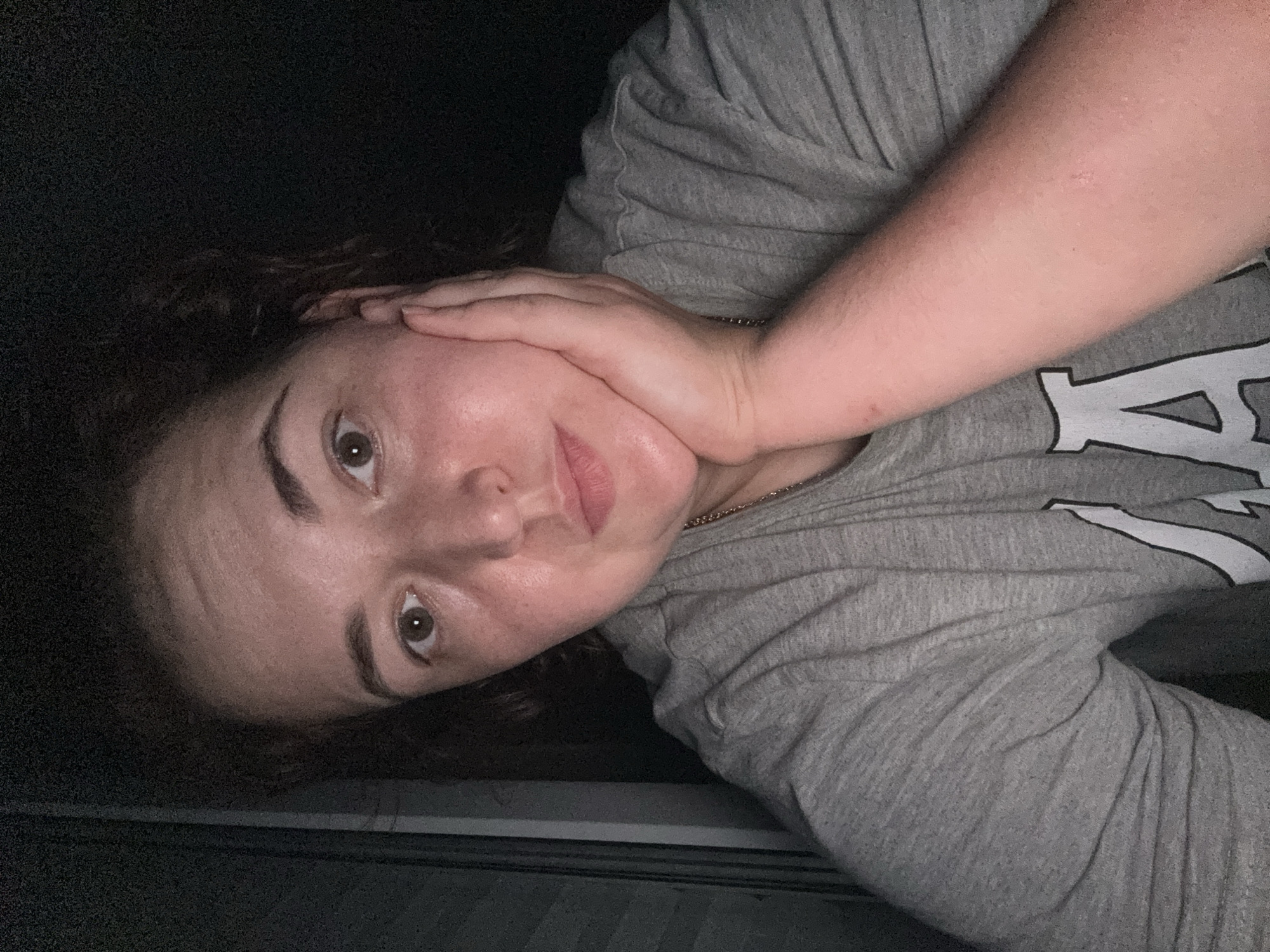 A selfie of a person is shown. The person's head is tilted slightly, and their right hand is resting on their chin. They are wearing a grey t-shirt. The background is dark, with some vertical lines visible on the left side, suggesting blinds or shutters. The image is dimly lit, casting shadows on the person's face and body. The location in Chelyabinsk, Russia, is not identifiable from the visual information in the image.