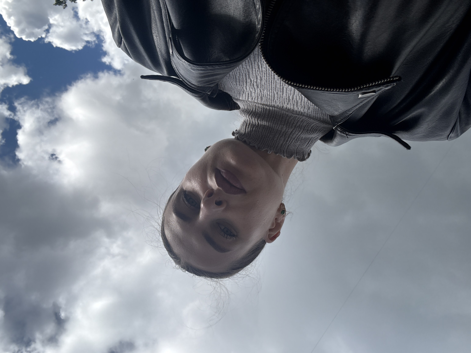 The image captures a person, likely female, from an extreme low-angle perspective, looking directly down into the camera. The subject is positioned against a bright, predominantly cloudy sky, with their head appearing inverted in the frame.
The person is wearing a black leather jacket, which is open to reveal a grey, ribbed, high-neck or turtleneck top underneath. A silver zipper and a small metal detail are visible on the jacket. A small, dark green stud earring is visible in the subject's right ear. Her hair is styled back, and her face shows a neutral to slightly contemplative expression, with her eyes focused downwards.
The background is an expansive sky, filled with a mix of white and grey clouds. In the upper left corner, patches of clear blue sky are visible, along with a hint of green tree foliage, indicating an outdoor setting. The lighting is natural and diffused, suggesting it is daytime. The image presents an unusual and artistic portrait. No specific activity beyond posing is evident. There is no visible text in the image. The scene is set in Chaikovsky, Russia, on a partially cloudy day.