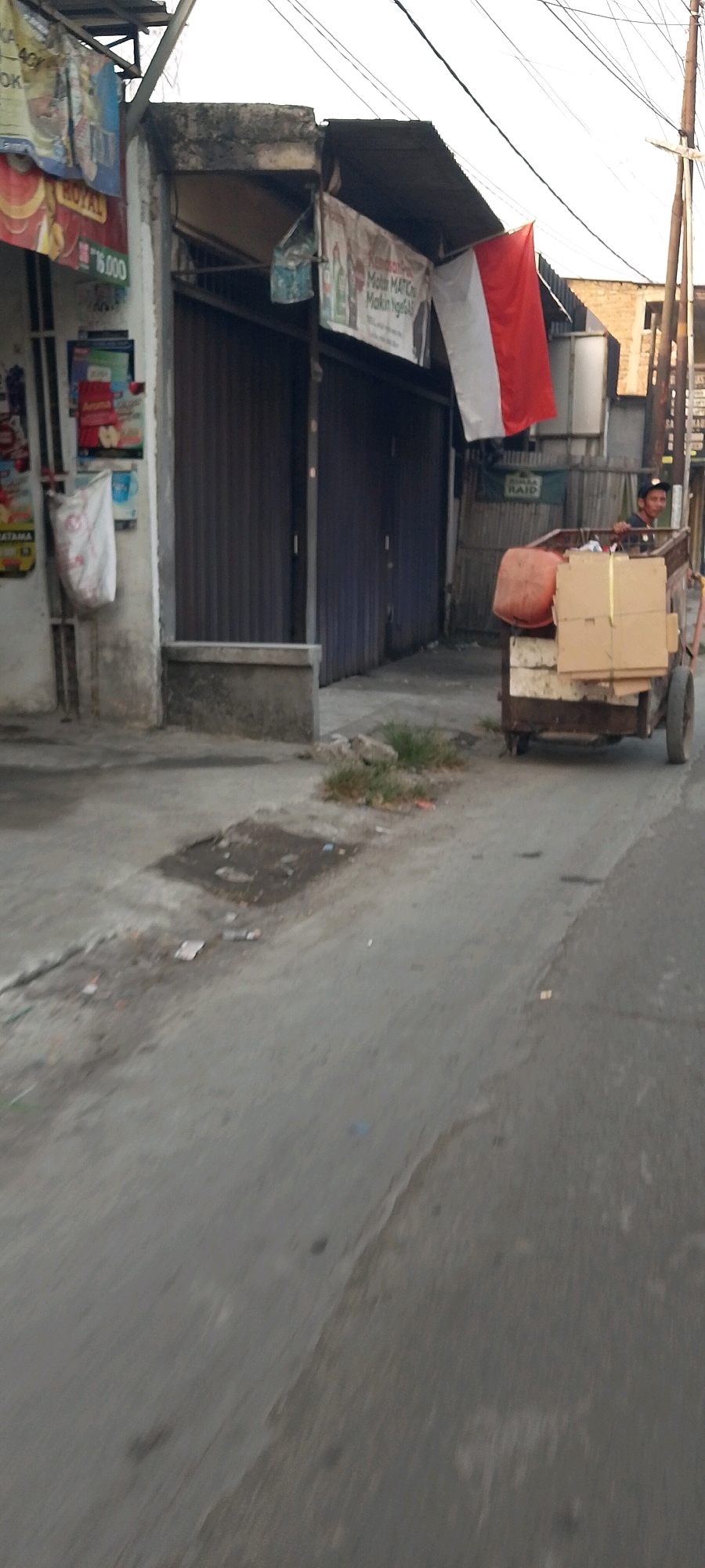 This image captures a street scene in Satriamekar, Indonesia. In the foreground, a worn asphalt road shows patches of discoloration and debris, including small pieces of trash and a darker, wet patch. To the left, a weathered concrete wall supports several posters and banners, one of which is white with red lettering. A white bag hangs from a hook on the wall.
Further down the road on the left, a building with a dark, ribbed metal shutter is visible. Above the shutter, a red and white flag, likely the Indonesian flag, hangs from the roofline. To the right of the flag, a banner with text and images is displayed.
Dominating the right side of the image is a hand-pulled cart laden with various items. A large orange cylindrical object, possibly a drum, is secured at the front of the cart. On top of the cart, several cardboard boxes are stacked and tied with yellow straps. A man wearing a dark shirt and a cap is seated on the cart, appearing to be operating or supervising its movement. In the background, more buildings and a utility pole are visible. The overall atmosphere suggests a typical urban environment with signs of commerce and daily life.