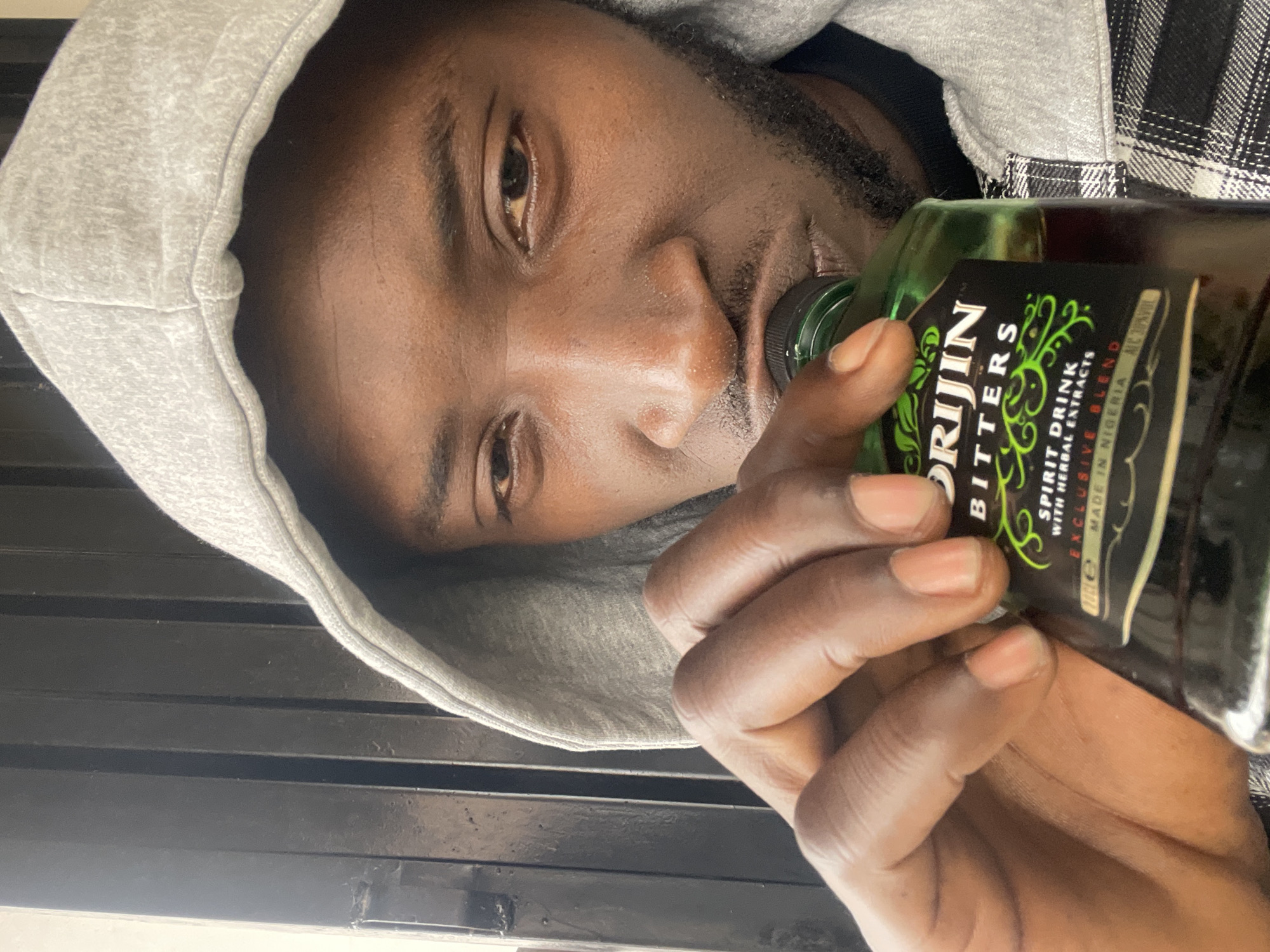 A man is holding and drinking from a bottle of "Orijin Bitters," a spirit drink with herbal extracts made in Nigeria. The man is wearing a grey hoodie. The location is unclear and not necessarily Jos, Nigeria, despite the drink's origin.
