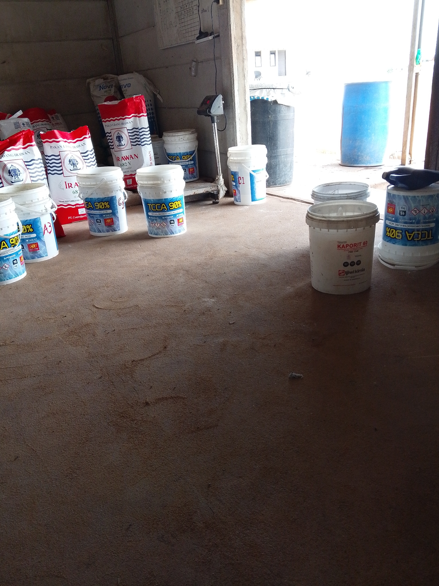 The image depicts a storage area in Bukit Ketok, Indonesia, likely a warehouse or outbuilding. In the foreground, there is a rough, reddish-brown floor with a few scattered debris. Several buckets and bags are distributed throughout the space. Bags labeled "PAKAN UDANG IRAWAN" (shrimp feed) and "Novo" are stacked against the back wall. Various buckets contain what appears to be chemical or treatment products, including "TCCA 90%" (Trichloroisocyanuric Acid) and "KAPORIT 60". There are also buckets labeled "CA 90%" (Cyanuric Acid) and "C1". In the background, there is a weighing scale with a digital display. Further back, past an open doorway, are blue and black plastic barrels. The scene appears to be a storage area for aquaculture or agricultural supplies.