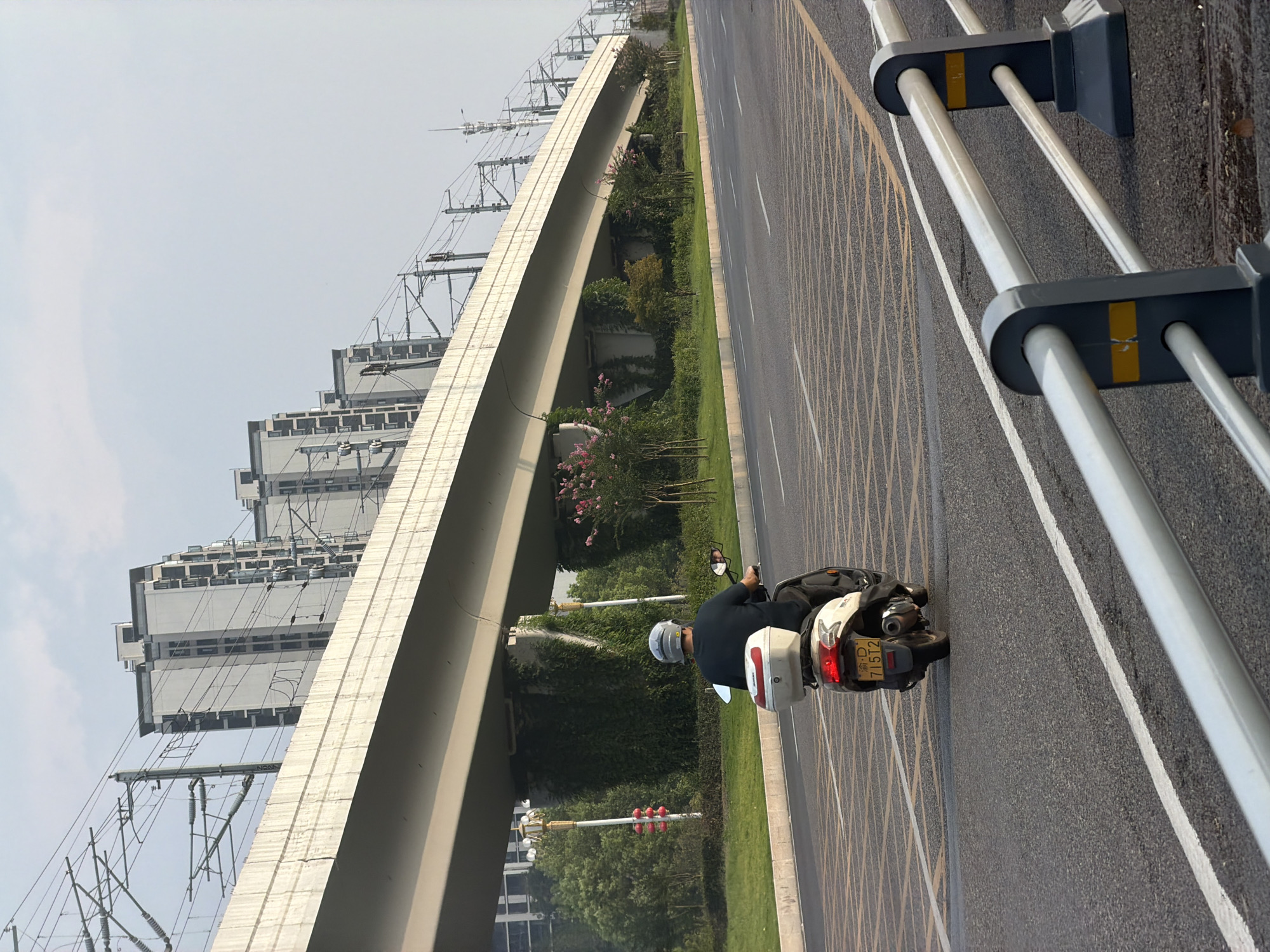 An overcast day in Jiangjin, China, features a person riding a white scooter away from the viewer on a multi-lane asphalt road. The scooter has a red and white storage box mounted on the back and a yellow license plate with the characters "D 715T2." The rider wears a dark shirt and a grey helmet. The road is marked with white lines and a yellow crosshatch pattern in the distance. A concrete overpass, possibly for a railway or elevated highway, stretches across the frame, supported by pillars draped in ivy. In the background, several tall, modern apartment buildings with dark windows stand against a hazy sky. Overhead, electrical wires and poles associated with the overpass are visible. On the left side of the road, a traffic light with three red lights is illuminated, and a decorative lamppost stands nearby. Green grass and scattered trees, some with pink blossoms, line the median and the embankment beneath the overpass. A gray metal guardrail with dark gray posts and yellow accents runs along the right side of the road in the foreground.
