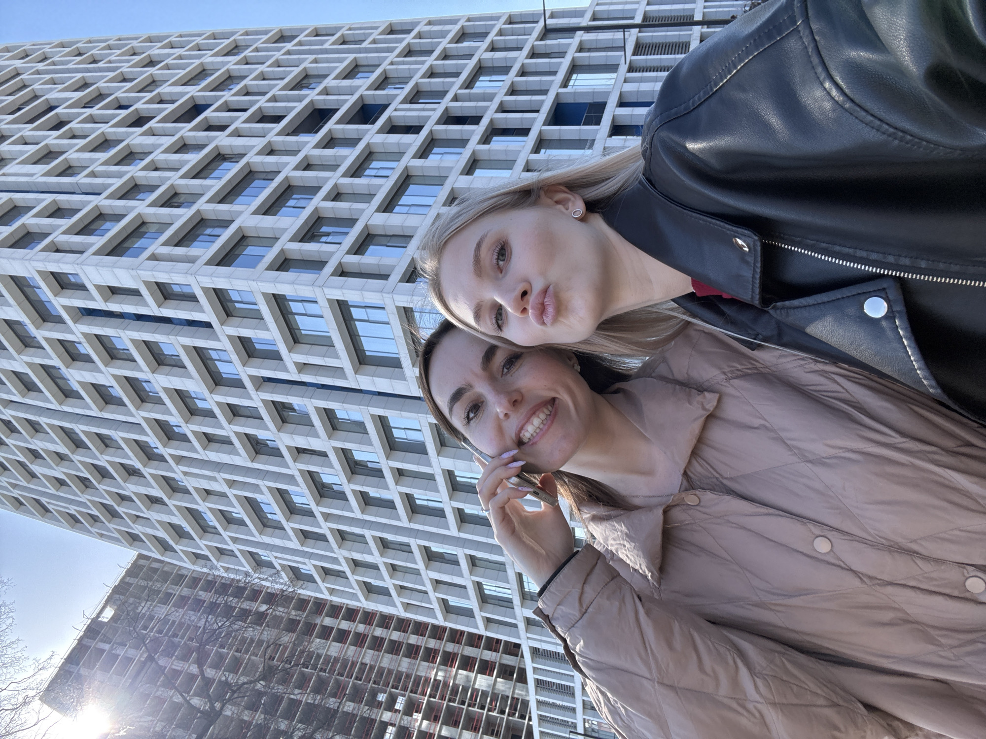 Two women are posing for a selfie with a large, modern building in the background. The blonde woman on the right is wearing a black leather jacket and puckering her lips. The brunette woman on the left is wearing a beige quilted jacket and holding a cell phone to her ear. Both women are smiling. The building in the background is white with a geometric pattern of windows. In the lower left of the frame, a second building is visible. The sky is clear and blue. The location is Izhevsk, Russia.