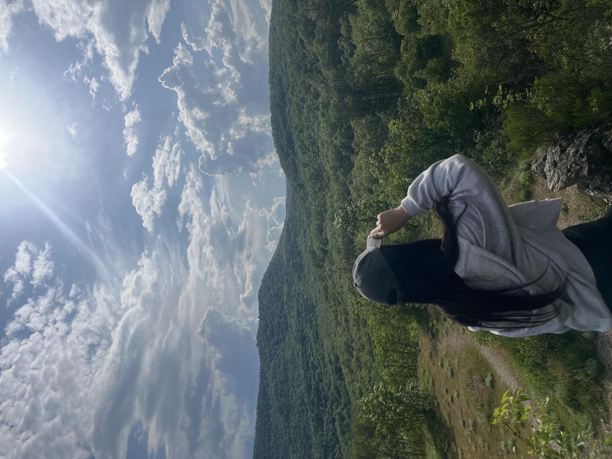 A medium shot captures a person in a gray hoodie and dark cap, standing at the edge of a grassy hill with a forested mountain as a backdrop. The sky is filled with dramatic, layered clouds, with the sun visible on the left side. The location appears to be in a natural, mountainous region, possibly near the city of Kamchia, Bulgaria, considering the landscape. The person's back is to the camera, and their arms are raised, possibly taking a picture. The scene has a rural and natural ambiance.