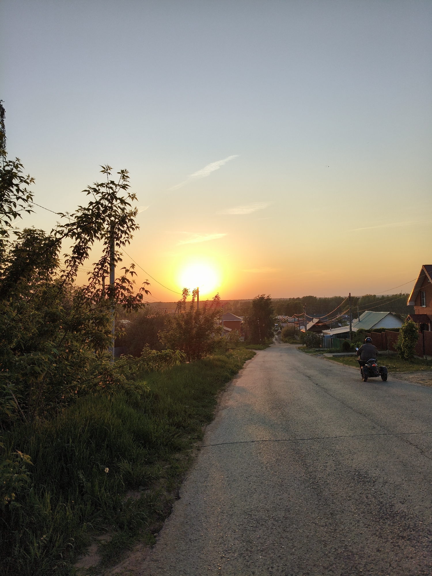 The photograph captures a scene at sunset in what appears to be a residential area in the city of Chaikovsky, Russia. The sun is setting over the horizon, casting a warm orange glow across the sky and landscape. A paved road stretches into the distance, and a three-wheeled vehicle with a rider is traveling along it towards the camera. On the left side of the road, there is lush vegetation and an electrical pole with wires. On the right, there are residential houses with visible roofs and fences. The overall atmosphere suggests a quiet, rural setting during the evening.