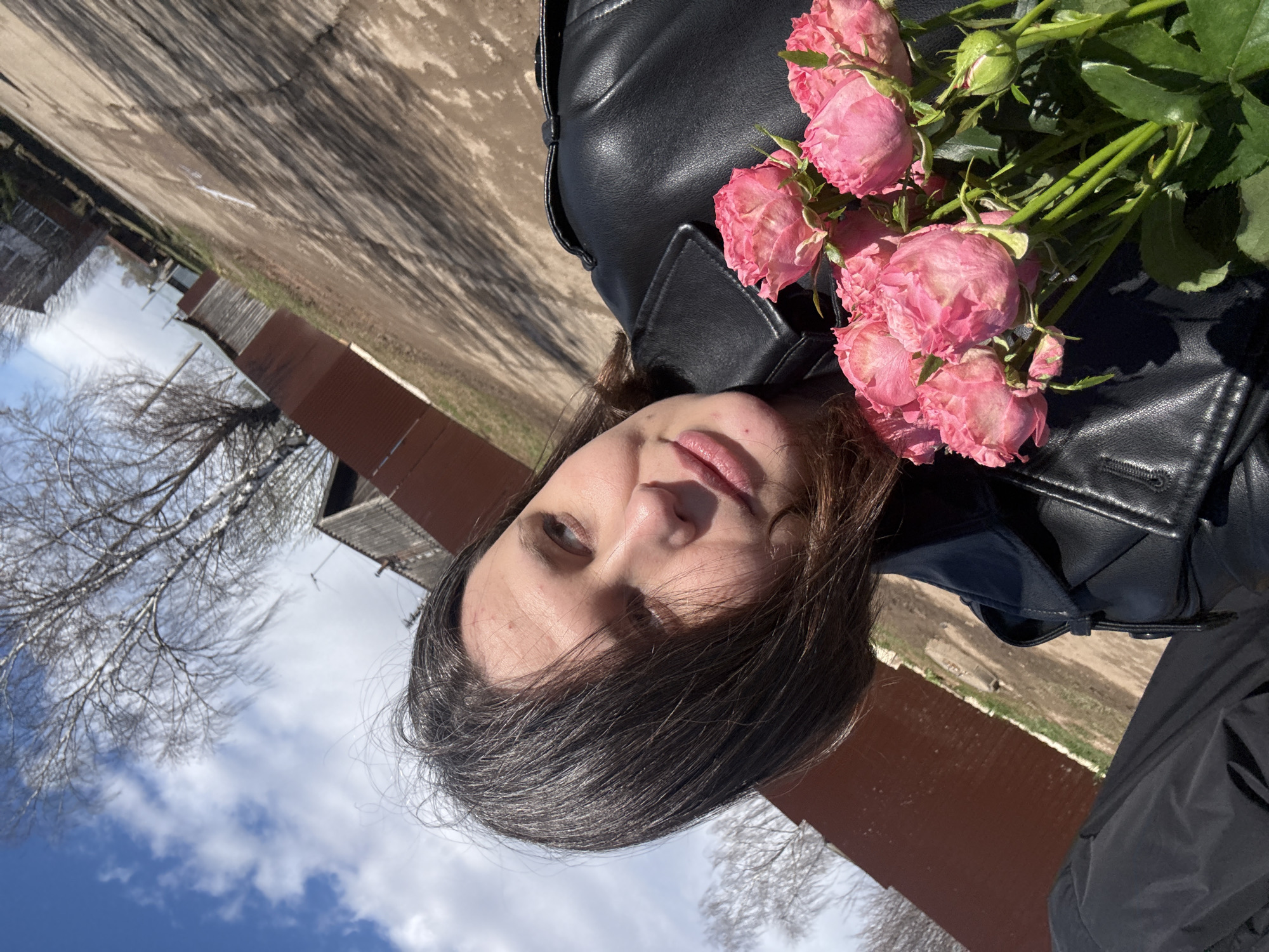 The image, rotated 90 degrees clockwise, depicts a woman posing outdoors with a bouquet of pink roses. She has medium-length dark hair, fair skin, and is wearing a black leather jacket. Her expression is neutral, and her eyes are looking slightly upward and to the side.
She holds a bunch of pink spray roses with green stems and leaves, positioned over her left shoulder and chest. The roses are in various stages of bloom, with some fully open and others as tight buds; some petals show slight signs of dryness or fading at the edges.
The background shows a bright blue sky with scattered white clouds. Several bare, leafless trees are visible, suggesting the time of year is late winter or early spring. In the middle ground, there's a structure with a reddish-brown corrugated metal wall or fence. The ground below appears to be dirt or dry grass, marked by long shadows from unseen objects, indicating strong daylight.
The activity is a posed photograph in an outdoor setting, likely in Фоки, Russia, given the context. The time of day is sunny daytime, and the weather is clear with some clouds. No text is visible in the image.
