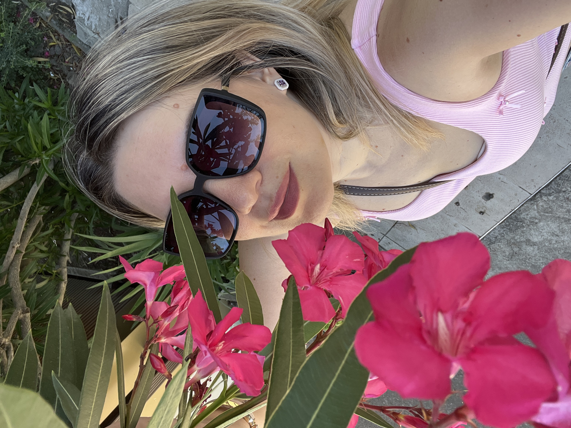 A woman is lying down outdoors, with pink flowers in the foreground. She has blonde hair with highlights and is wearing large, dark sunglasses. Her reflection in the sunglasses shows foliage. She is also wearing a pink ribbed tank top with a small bow detail. A single earring with a purple gem is visible in her ear. The background consists of greenery and a gray, paved surface. The overall impression is that of a casual, sunny day, possibly in a garden or park. The flowers suggest a warm climate.