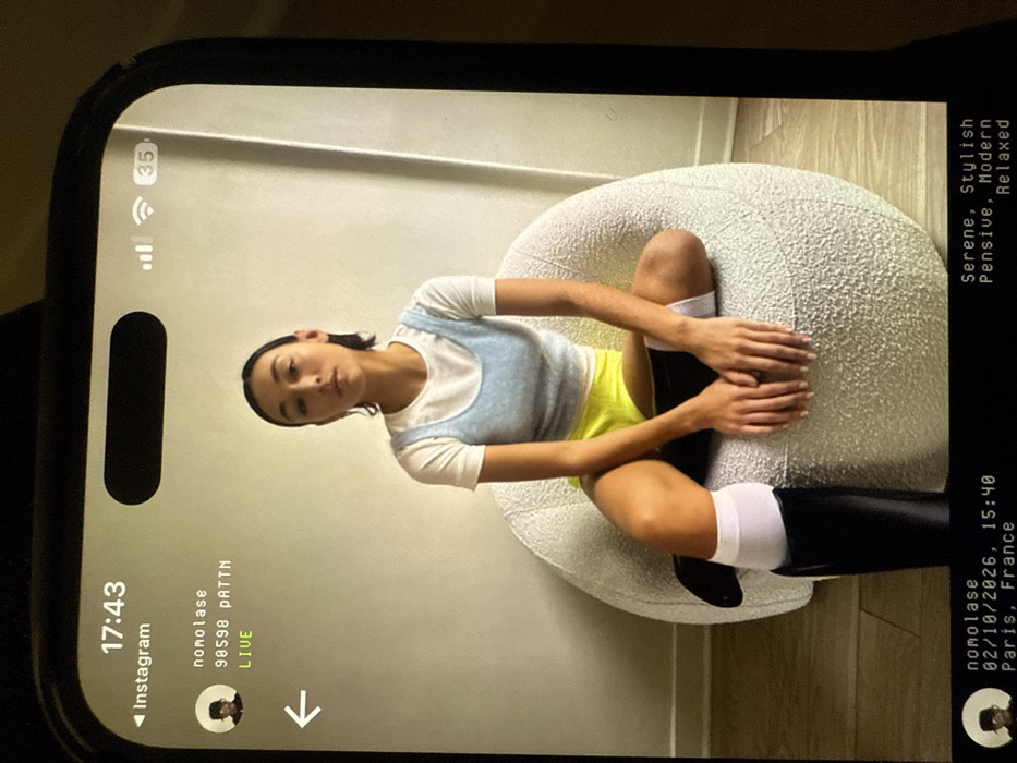 A phone screen displays an Instagram Live stream of "nomolase." The live video features a woman sitting cross-legged on a textured, round chair. She wears a white t-shirt under a light blue knit top, neon yellow shorts, and black knee-high socks with white cuffs. Text overlay on the right side of the screen reads "Serene, Stylish, Pensive, Modern, Relaxed" and "02/10/2026, 15:40 Paris, France." The upper left corner shows the time "17:43" and a cellular signal. A download arrow is displayed.