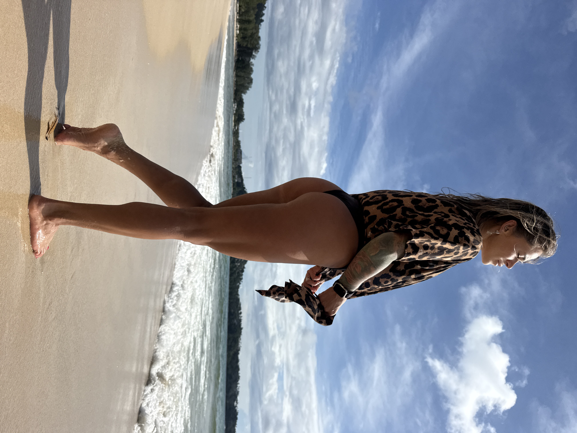 A young woman stands on a sandy beach in Choeng Thale, Thailand, facing right with her head turned to look out at the ocean. She has blonde hair that is wet and pulled back, and she is wearing a leopard-print short-sleeved shirt over a black bikini bottom. Her left arm is bent, and she is holding the shirt with her hands, one of which has a watch on her wrist. She has tattoos on her left arm and shoulder. The beach is light brown sand, and the ocean is a light blue-green with white waves breaking on the shore. In the background, there is a line of green trees and buildings along the coast. The sky is blue with scattered white clouds. The sun is shining, casting a shadow from the woman on the sand.