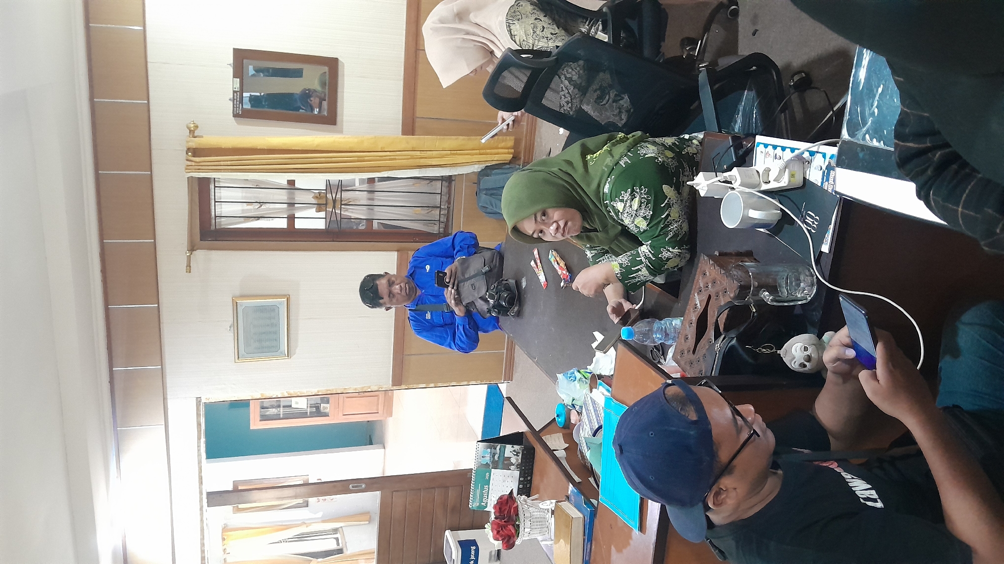 In a room that appears to be an office in Blora, Indonesia, four individuals are gathered around a dark brown table. A man in a blue shirt stands, engrossed in his smartphone, with a grey backpack and a black camera on the table before him. To his right, a woman wearing a green hijab and a floral patterned dress looks towards the camera, holding a smartphone. Across from her, another person, partially obscured, is also looking at a smartphone. In the foreground, a man wearing a black t-shirt and a blue baseball cap is also focused on his smartphone, which is plugged into a charger on the table. The table is cluttered with various items, including a tissue box, a glass, a white mug, a water bottle, and papers. The room has light-colored walls and a wooden ceiling with inset panels. A doorway with a glass-paneled door is visible in the background, as is a window with yellow curtains. The overall impression is one of a casual meeting or work session where individuals are concurrently engaged with their mobile devices.