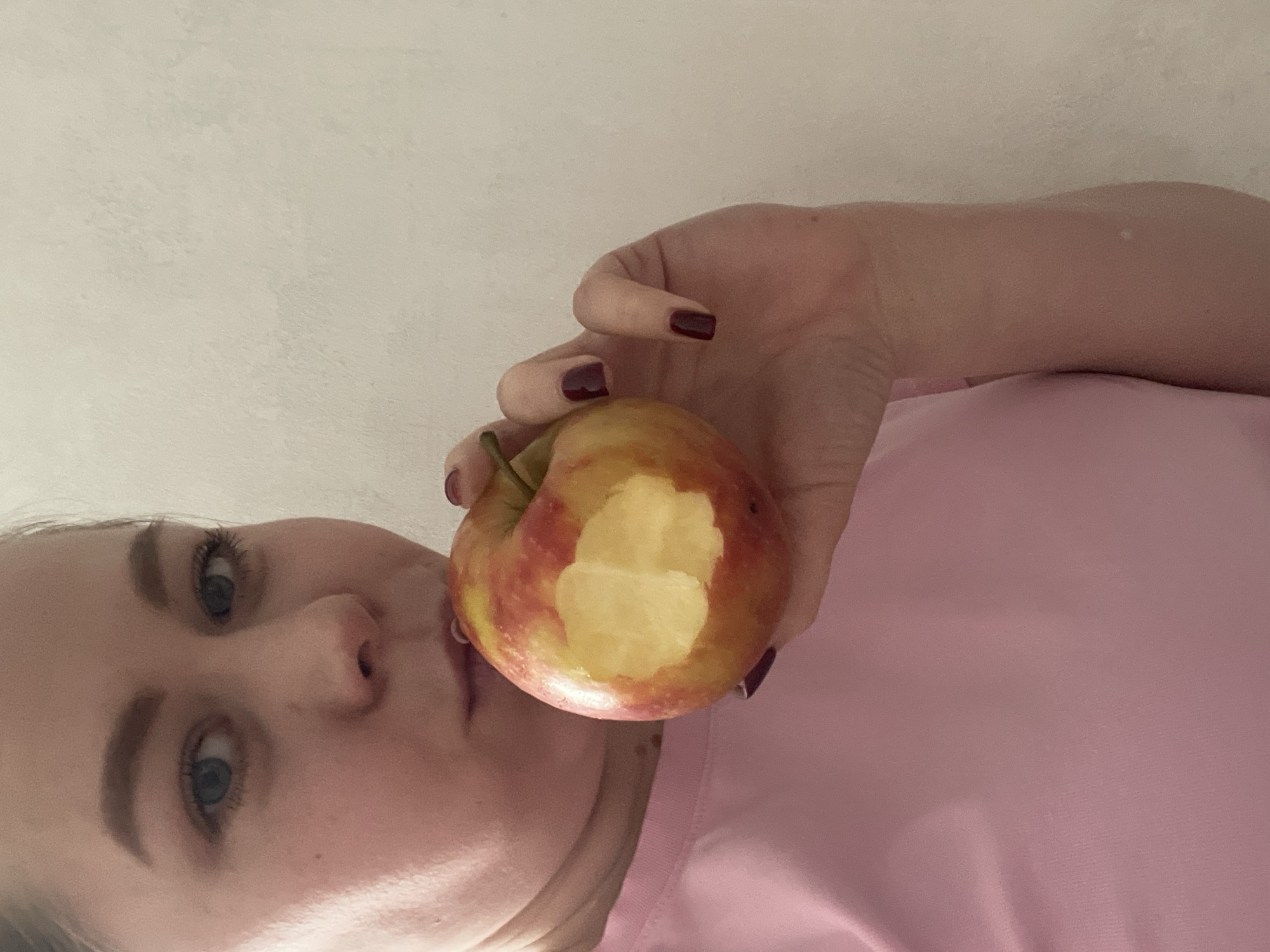 A woman is holding a partially eaten apple close to her face. The apple is red and yellow, with a bite taken out of it. The woman is wearing a pink shirt and has dark red nail polish. The background is a plain white wall. There is no indication of Chaikovsky, Russia, in the image.