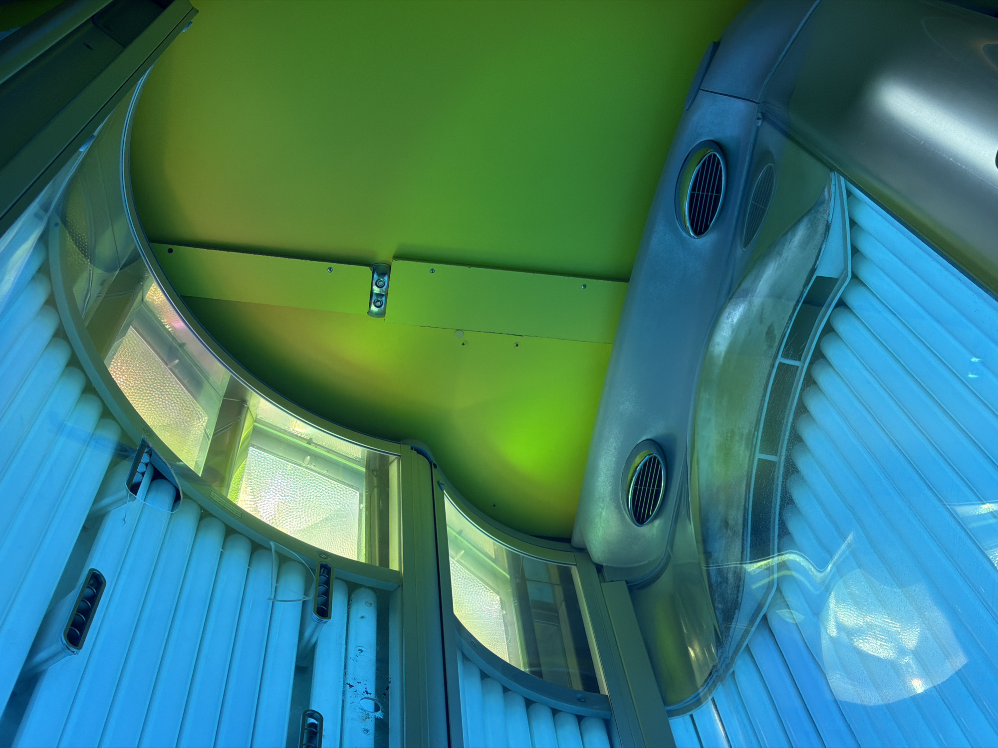 An interior view of a tanning bed with bright blue and green lighting. The tanning bed features a series of fluorescent tubes that emit blue light, illuminating the ribbed surface of the bed. Above, a curved, green wall is visible, with metal fixtures and a ribbed vent. A clear, curved acrylic surface reflects the blue light and a textured, light-colored surface beneath. The image is devoid of any people or human activity and does not contain any contextual clues to indicate its location in Kyiv, Ukraine.