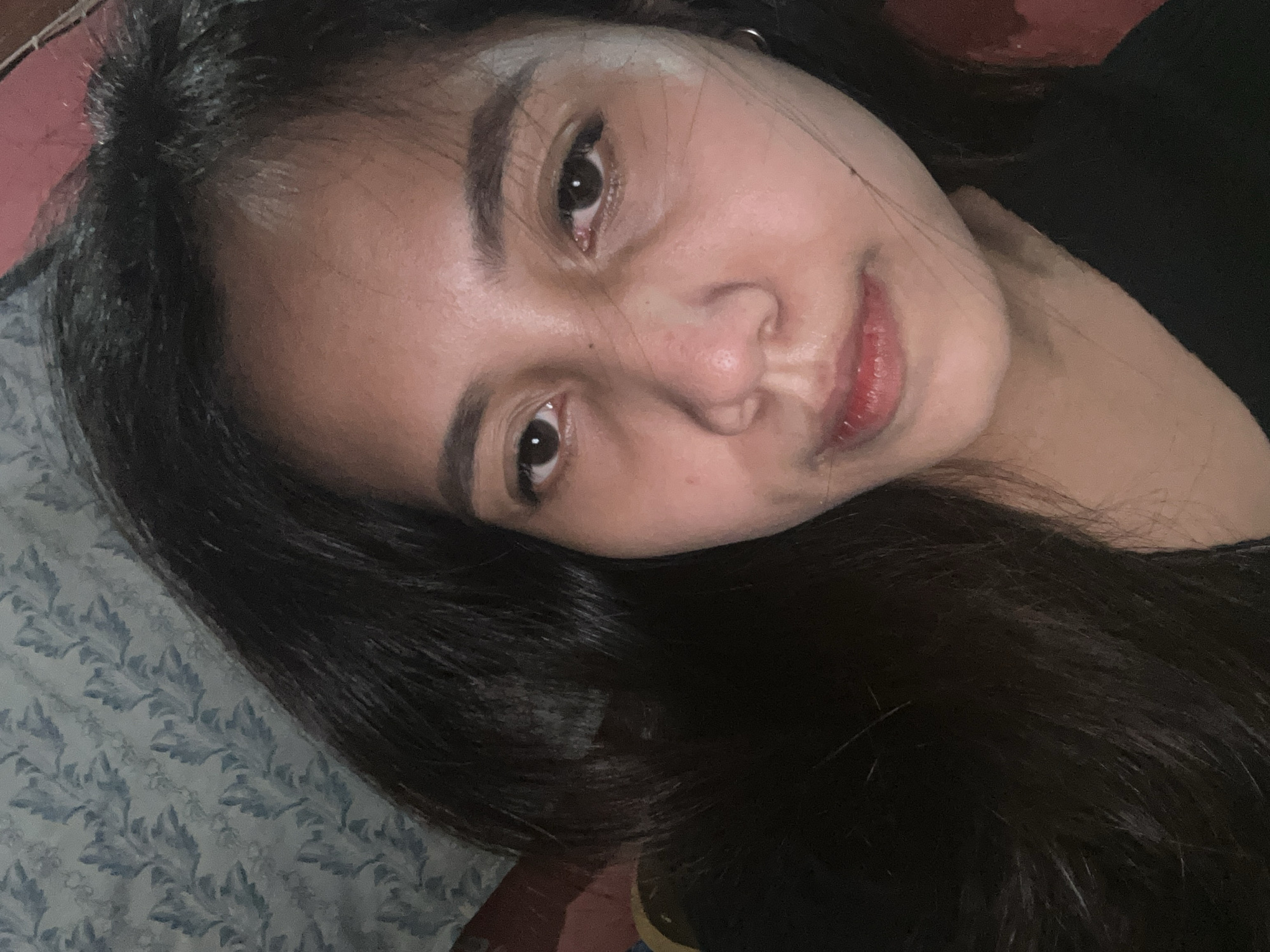 The image is a close-up selfie of a young woman. She has dark hair that partially covers her face. Her skin appears to be fair to medium toned, with some shine visible, possibly from sweat or makeup. Her eyes are dark brown, and her eyebrows are well-defined. Her lips are painted with a reddish-pink lipstick. She is wearing a black top. The background is out of focus but appears to be a wall with a patterned wallpaper. A small hoop earring is visible in her left ear. The lighting suggests an indoor setting, possibly a bedroom, given the soft light and the relaxed pose. There is no information in the image to indicate its specific location within the city of Depok, Indonesia.