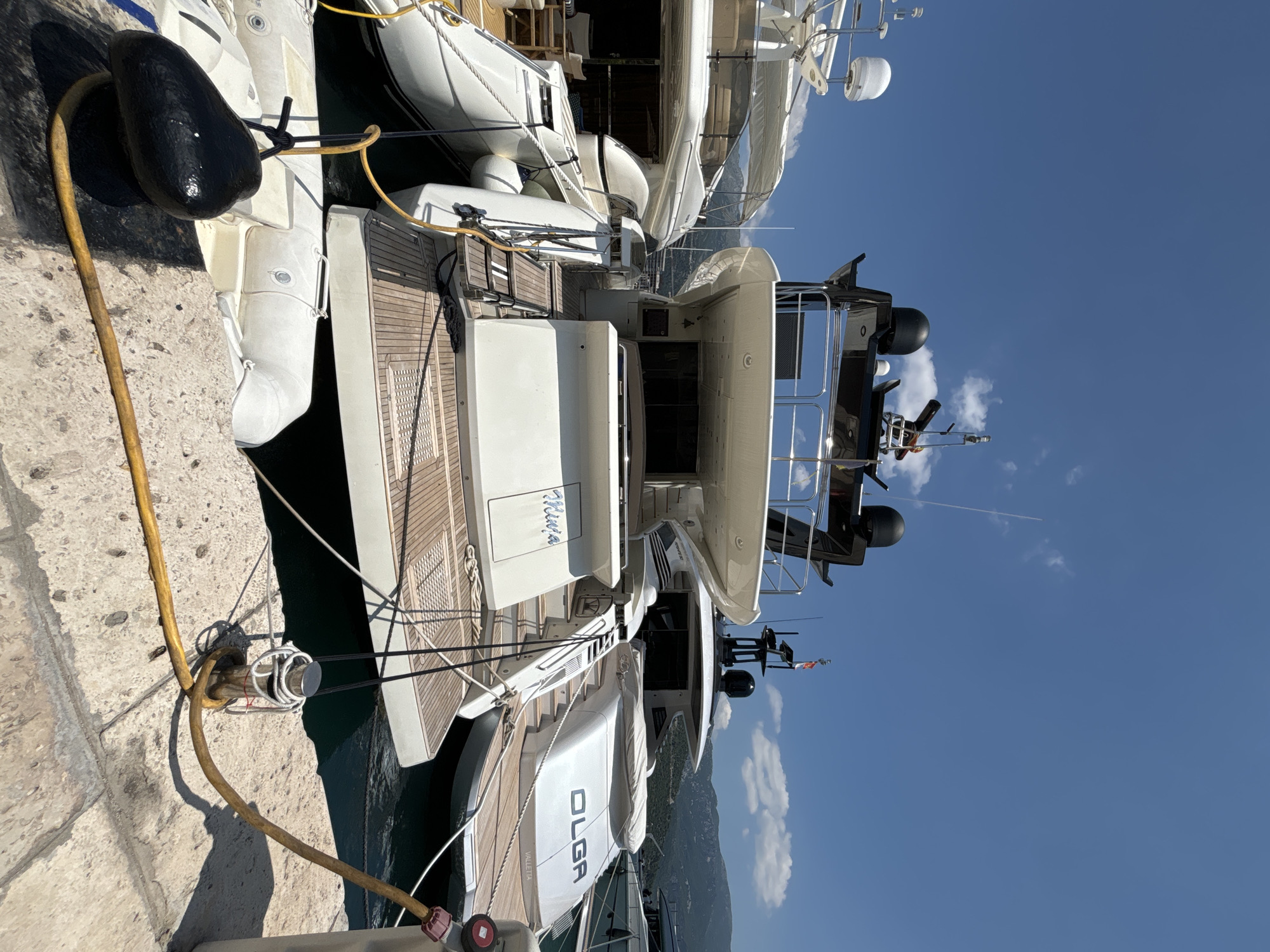 This media file shows a large white yacht docked at a pier in Budva, Montenegro. The yacht is detailed with wooden decking and several white inflatable tenders. One tender is clearly labeled "OLGA VALLETTA." A black, oblong fender is attached to the pier by a yellow rope and a black rope. The scene is bathed in bright sunlight, indicating it is daytime. The sky is a clear blue with a few scattered white clouds. In the background, mountains rise steeply, their slopes covered in greenery. The overall impression is one of a peaceful and luxurious maritime setting.