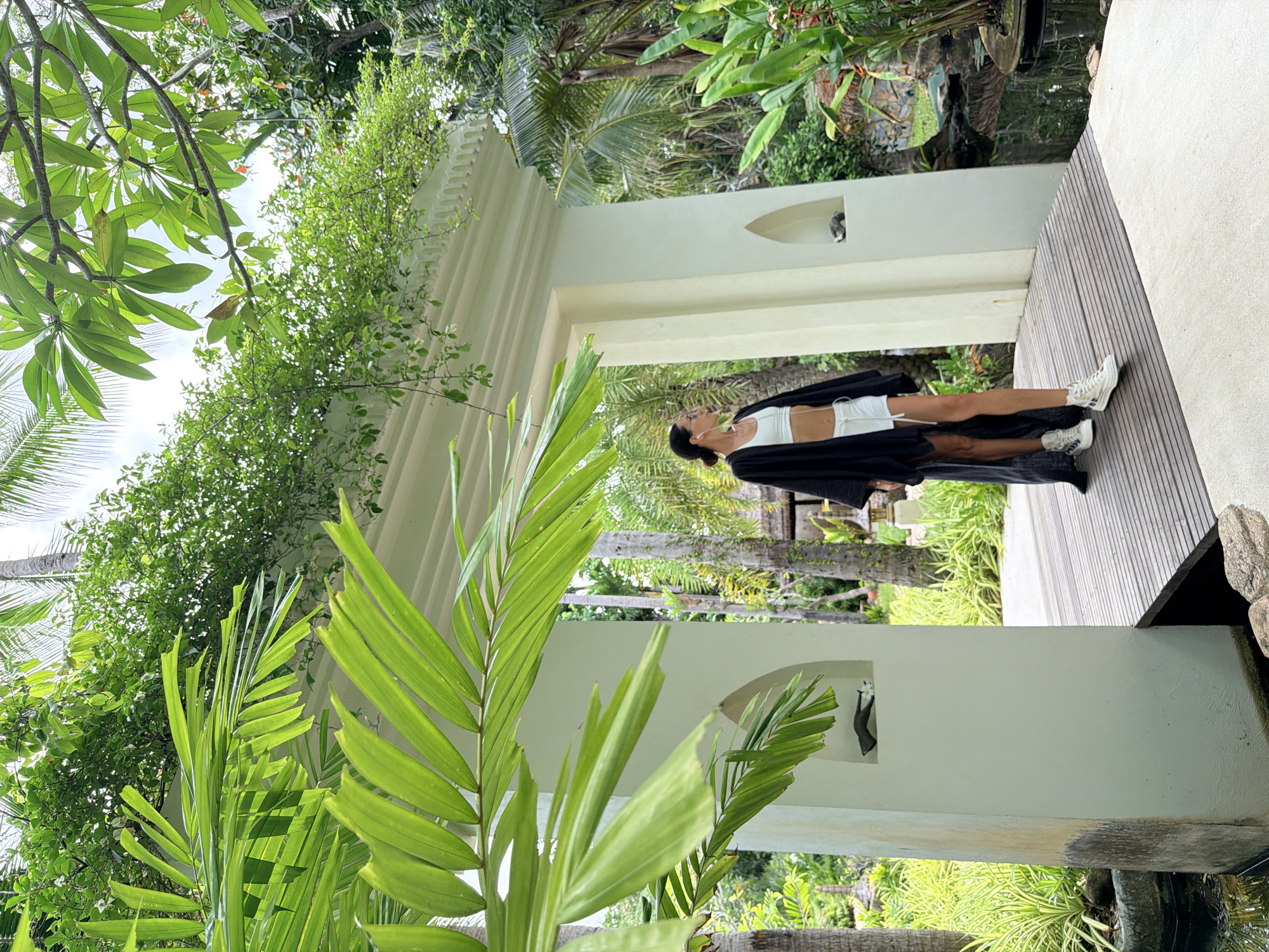 In the city of Ko Samui District, Thailand, a woman stands on a wooden bridge, her dark kimono flowing around her. She wears a white crop top and white shorts, with white high-top sneakers on her feet. Her hair is pulled back, and she wears earrings. The bridge leads through a white archway adorned with decorative carvings. Lush green foliage surrounds the area, with palm trees visible in the background. A small pond is situated to the right of the bridge, with rocks and plants bordering it. The environment suggests a tranquil, tropical setting, possibly a resort or a private garden.