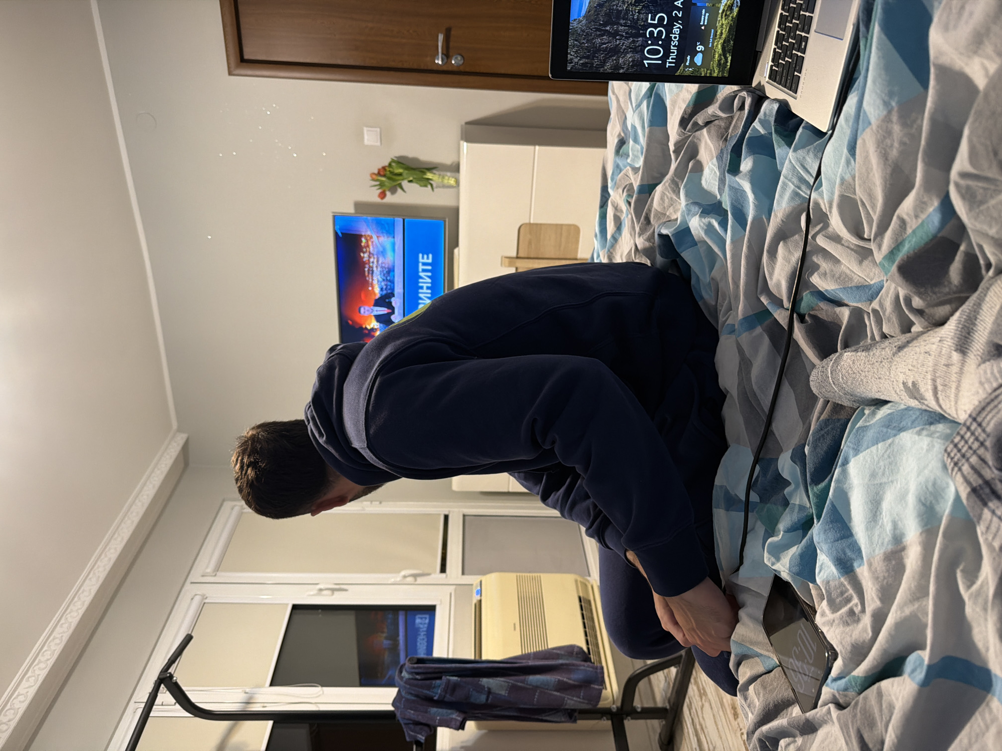 A man in a navy blue tracksuit is reaching into a bed covered with blue, white, and grey patterned sheets. A laptop sits on the bed next to him, its screen displaying "10:35, Thursday, 2 August, 9°C." The man is holding a mobile phone in his left hand. A power cord snakes from the laptop towards the phone. In the background, there is a television showing a news program with Cyrillic text on the screen. Above the television, there is a vase of tulips. Further in the background, a door and a window are visible, along with a stationary bicycle. The setting is an interior room, possibly a bedroom. It is not possible to determine the exact location within Plovdiv, Bulgaria.