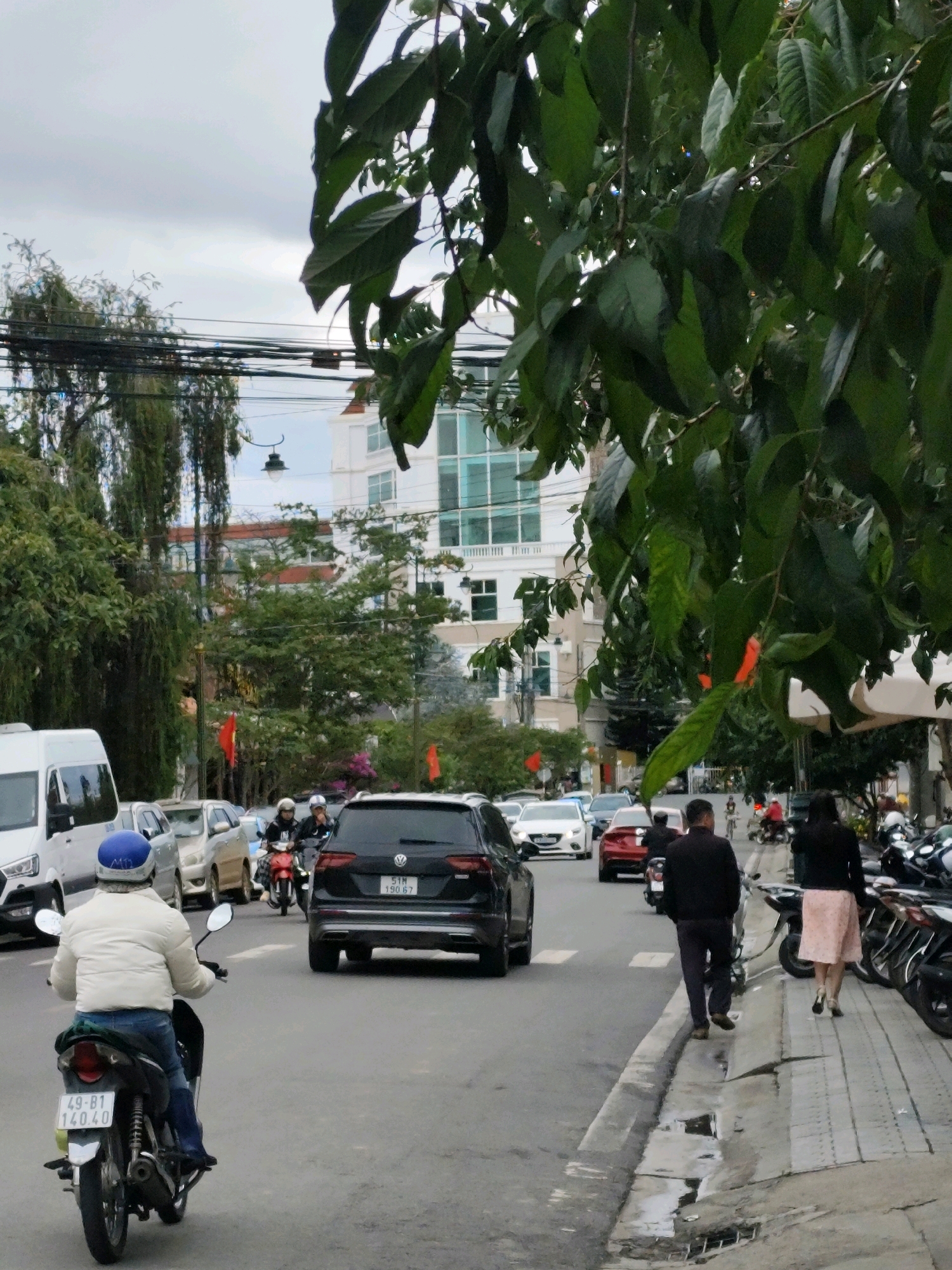 The image depicts a street scene in Lâm Viên - Đà Lạt, Vietnam, characterized by a mix of vehicles and pedestrians. In the foreground, a person wearing a white jacket and blue jeans rides a motorcycle with a blue helmet. The motorcycle has a license plate reading "49-B1 140.40". To the right of the motorcycle, a black SUV with a license plate "51H 190.67" is visible. Further down the street, a man in a dark jacket and dark pants walks away from the camera, while a woman in a pink skirt and black top walks beside him. Several other motorcycles are parked on the right side of the street. The street is lined with buildings of varying architectural styles, including a white building with glass windows and a building with red accents. Trees with lush green foliage partially obscure the view of the buildings and the sky. The sky is overcast, and the overall lighting suggests a cloudy day. Red flags are visible on poles along the street. The road surface shows signs of wear, and there are puddles on the sidewalk. The scene conveys a sense of everyday urban activity in Đà Lạt.