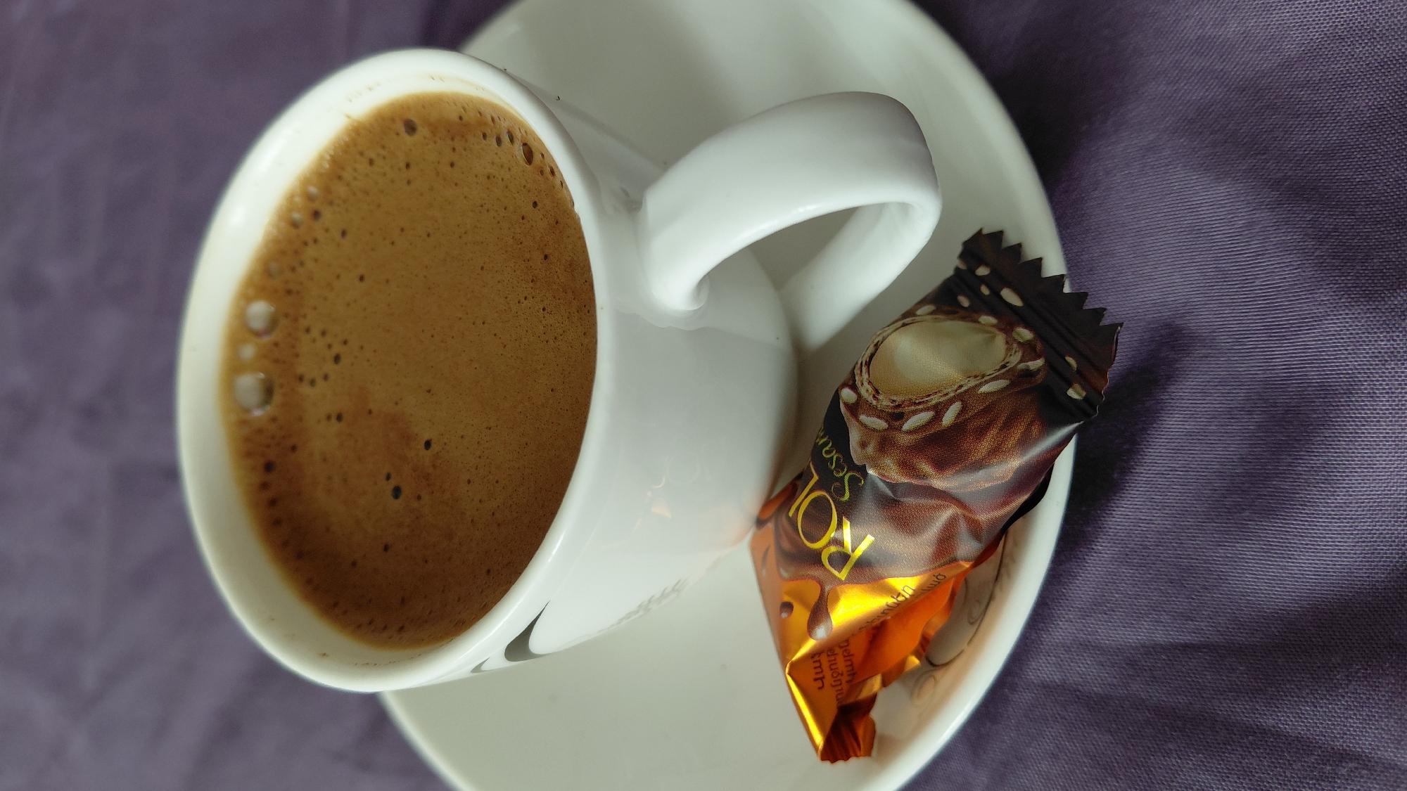 A close-up, overhead shot features a white ceramic cup filled with dark, frothy coffee. The cup rests on a white saucer, which is positioned on a textured, purple fabric surface. In front of the cup, on the saucer, lies a foil-wrapped chocolate candy. The candy wrapper is predominantly orange and brown, with visible text and an image of the chocolate itself, which appears to be filled with a creamy white center and topped with nuts or seeds. The lighting suggests an indoor setting, with soft shadows cast by the cup and candy. There are no people visible in the image. The scene is likely a casual moment, possibly a coffee break, in Abovyan, Armenia, given the context provided.
