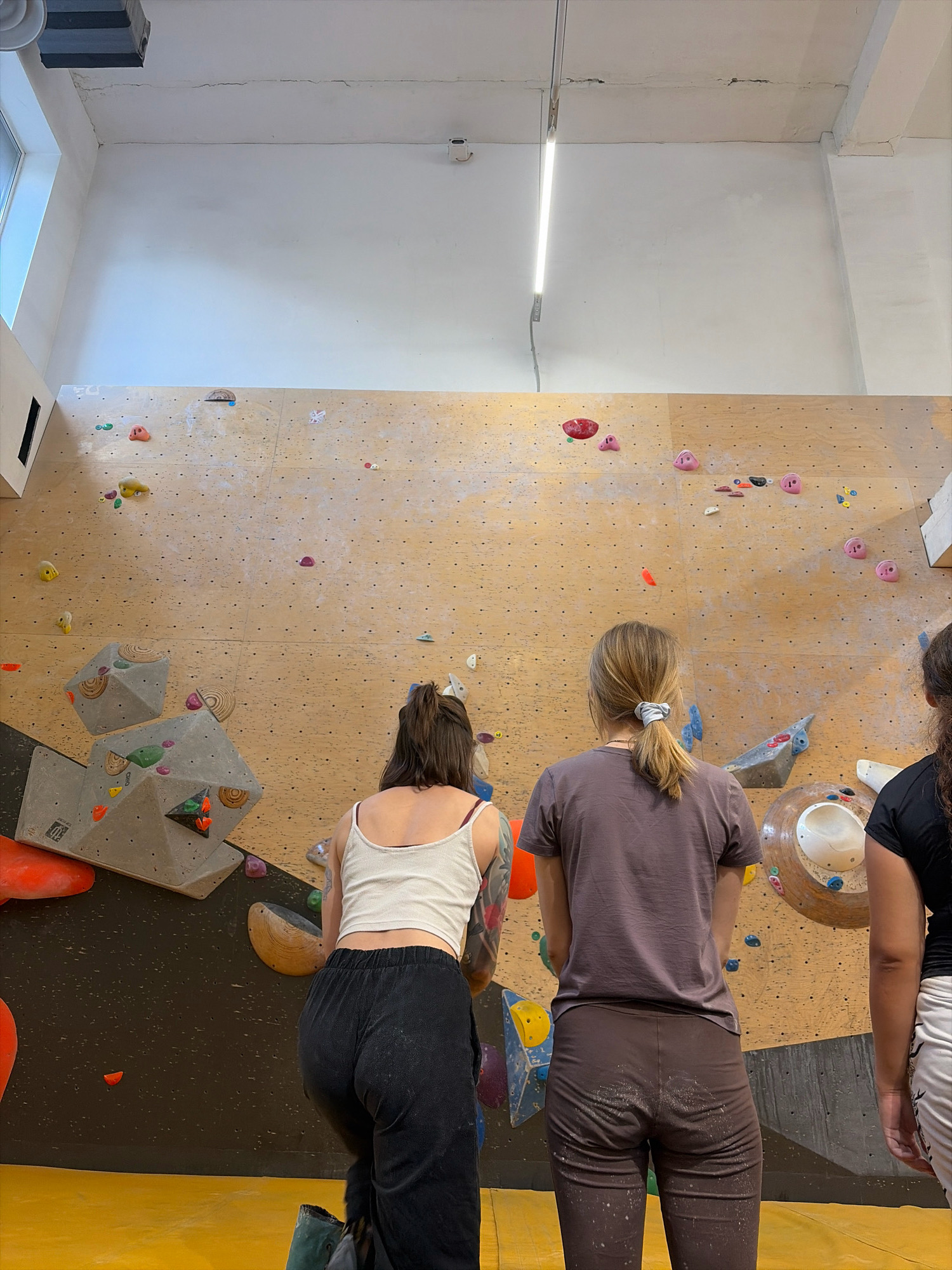 The image depicts three individuals, viewed from behind, standing in front of a bouldering wall. The wall is constructed from plywood with numerous small holes and is adorned with various climbing holds of different colors and shapes, suggesting it is part of an indoor climbing gym. Two of the individuals are closer to the foreground, facing the wall, while a third person is partially visible on the right. The environment appears to be well-lit, with overhead lights and natural light from a window visible on the left side of the frame. The floor is yellow. The scene is not identifiable as being in Moscow, Russia.