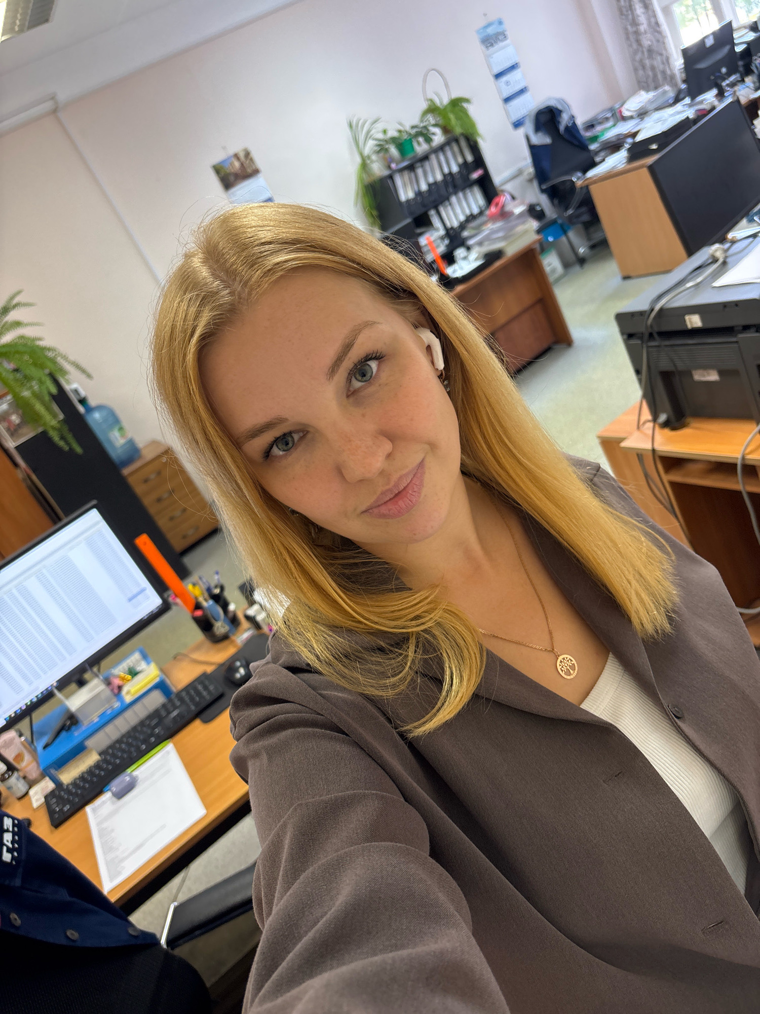 The media file is a selfie taken by a young woman in an office setting. She has blonde hair and is wearing a grey blazer over a white top. She has on white wireless earbuds and a necklace with a tree of life pendant. The background shows a cluttered office with desks, computers, printers, shelves filled with binders, and plants. The overall impression is that she is at work, possibly taking a break or a quick photo opportunity. There is no specific indication of the time of day or weather conditions, but the lighting suggests it might be daytime. The location is identified as Rusanovo, Russia. Visible text includes what appears to be a label on her dark blue shirt at the bottom left of the image, reading "ГАЗ".