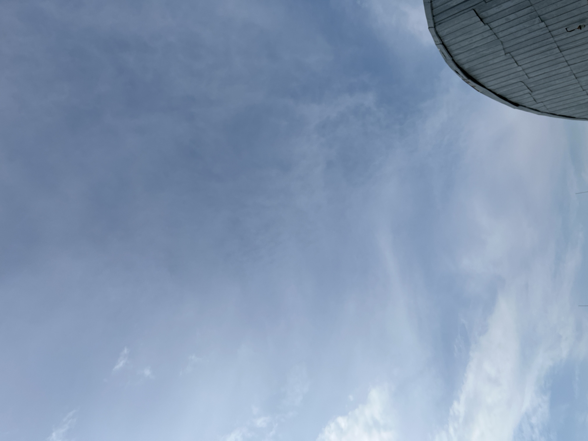 The image captures a portion of a domed structure, likely a roof, against a pale blue sky filled with wispy clouds. The structure appears to be made of metal panels arranged in a grid pattern, with a curved edge visible at the top right of the frame. The sky is overcast with thin, diffused clouds, suggesting a daytime scene with possibly mild weather. There are no people or animals visible in the image. The perspective is from below, looking upwards. The location context is Malyi Oleksyn, Ukraine, though this is not visually evident from the image itself. No text is visible.