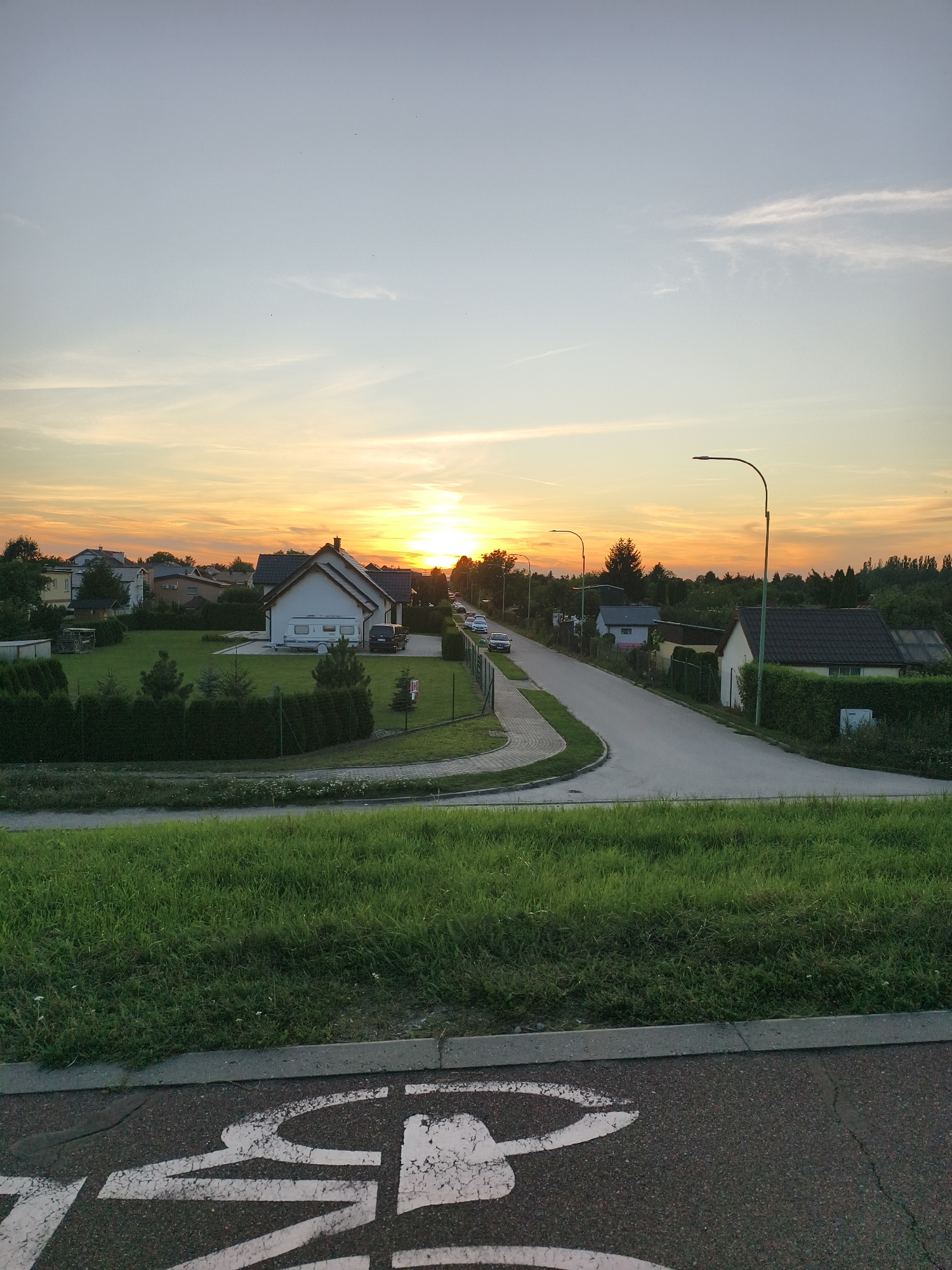 This is a photograph taken during sunset in a residential area, likely on the outskirts of Tczew, Poland. The sky is a gradient of pale blue and warm orange, with thin clouds streaking horizontally across the upper portion of the image. The sun is setting on the right side of the frame, casting a golden glow over the scene.
In the middle ground, a paved road curves through a neighborhood of houses. Several houses are visible, featuring pitched roofs and manicured lawns. A white caravan is parked in the driveway of one house. Cars are parked along the side of the road, and streetlights are positioned along the pavement. Trees and hedges line the properties, and a fence encloses some of the yards.
The foreground features a grassy embankment with a paved path or road running along its base. A white bicycle symbol is painted on the dark asphalt surface, indicating a bike lane. The overall impression is one of a peaceful evening in a suburban setting.