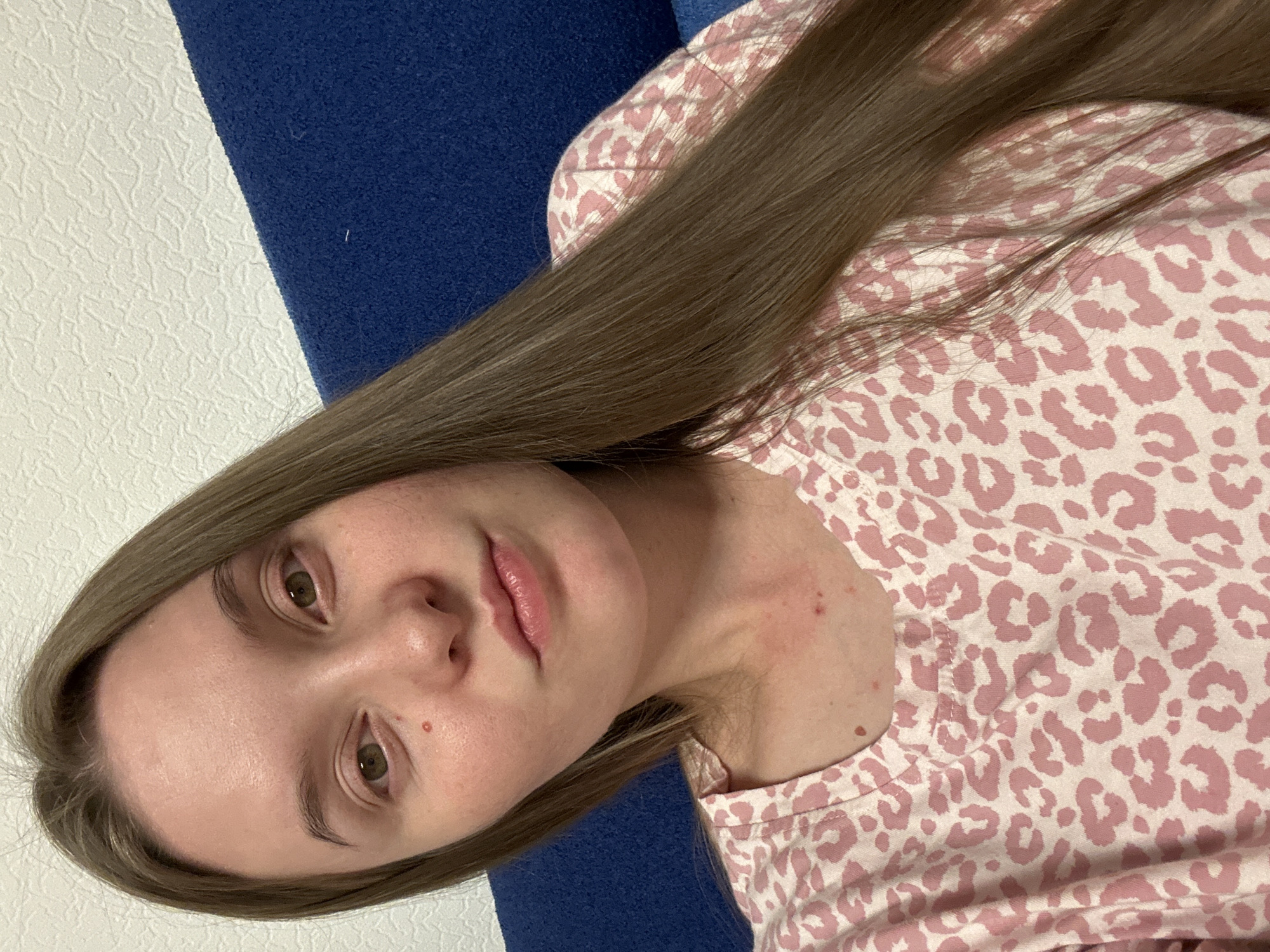 A young woman with long, straight brown hair is pictured from the chest up, seated against a textured white wall and a blue upholstered surface. She is wearing a pink and white leopard print shirt. Her expression is neutral, and her gaze is directed towards the camera. The lighting is soft, and the overall impression is one of a casual, indoor setting. There is no information about the location in Noviy, Russia, or any forensic details that can be extracted from the image.