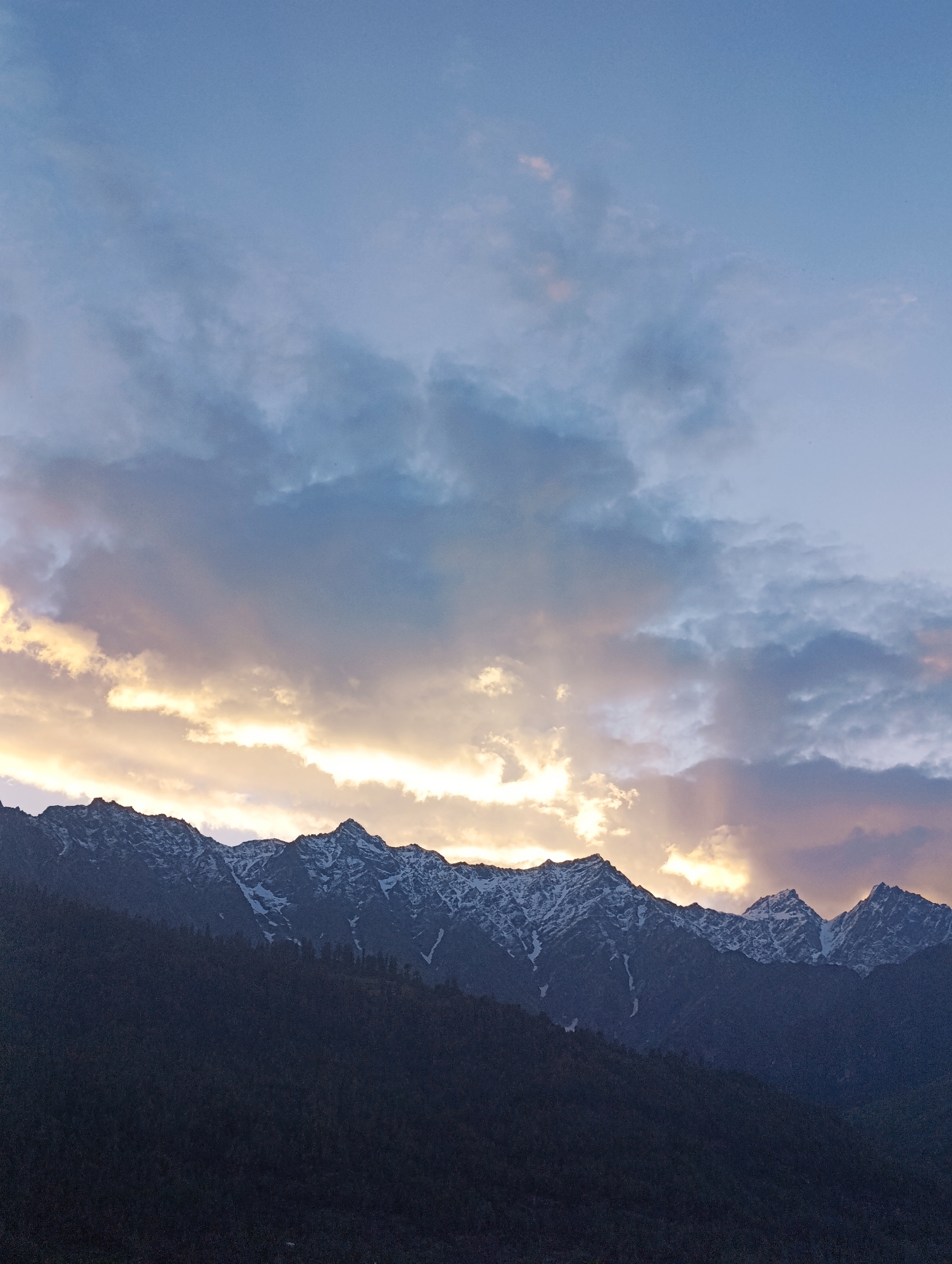 A landscape photograph captures a mountain range at dusk. The sky is filled with dramatic clouds, with patches of golden light breaking through, suggesting the last rays of the setting sun. The peaks of the mountains are snow-capped, contrasting with the dark, forested slopes below. The overall impression is one of serene natural beauty. The location is Vashisht, India.