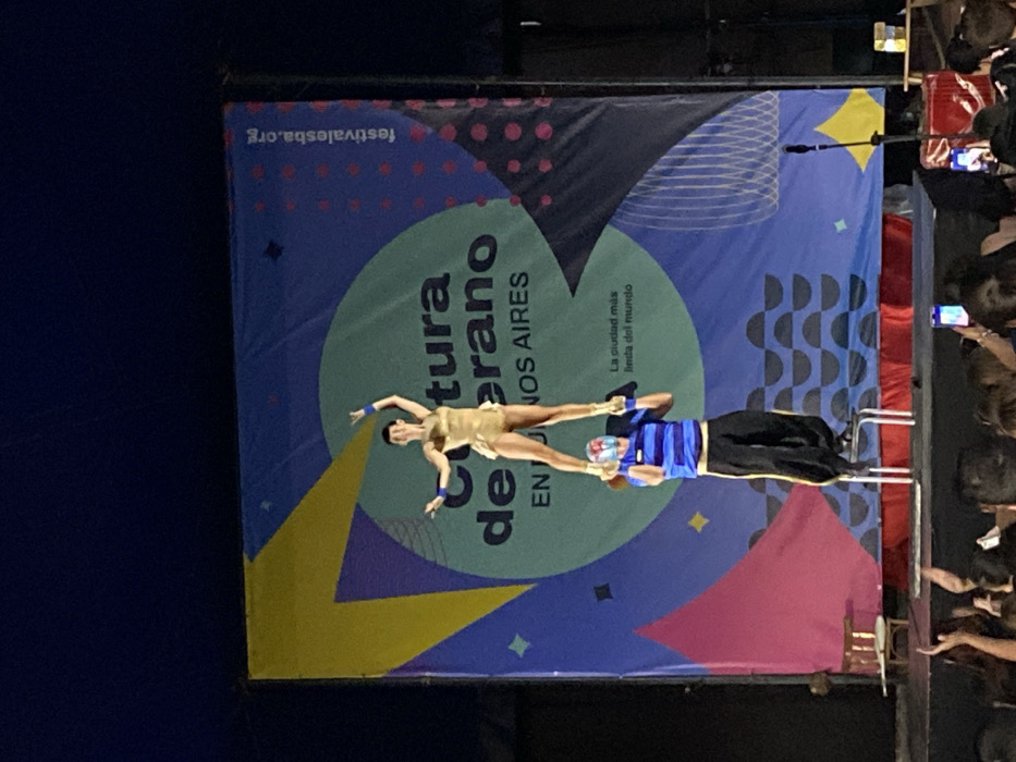 Two acrobats perform on stage in front of a banner promoting "Cultura de Verano en Buenos Aires". One acrobat, dressed in gold, balances atop the other, who is dressed in blue and black. The crowd is partially visible to the right of the performers, many holding up phones. A microphone stands to the left. The backdrop is colorful with geometric shapes and text.