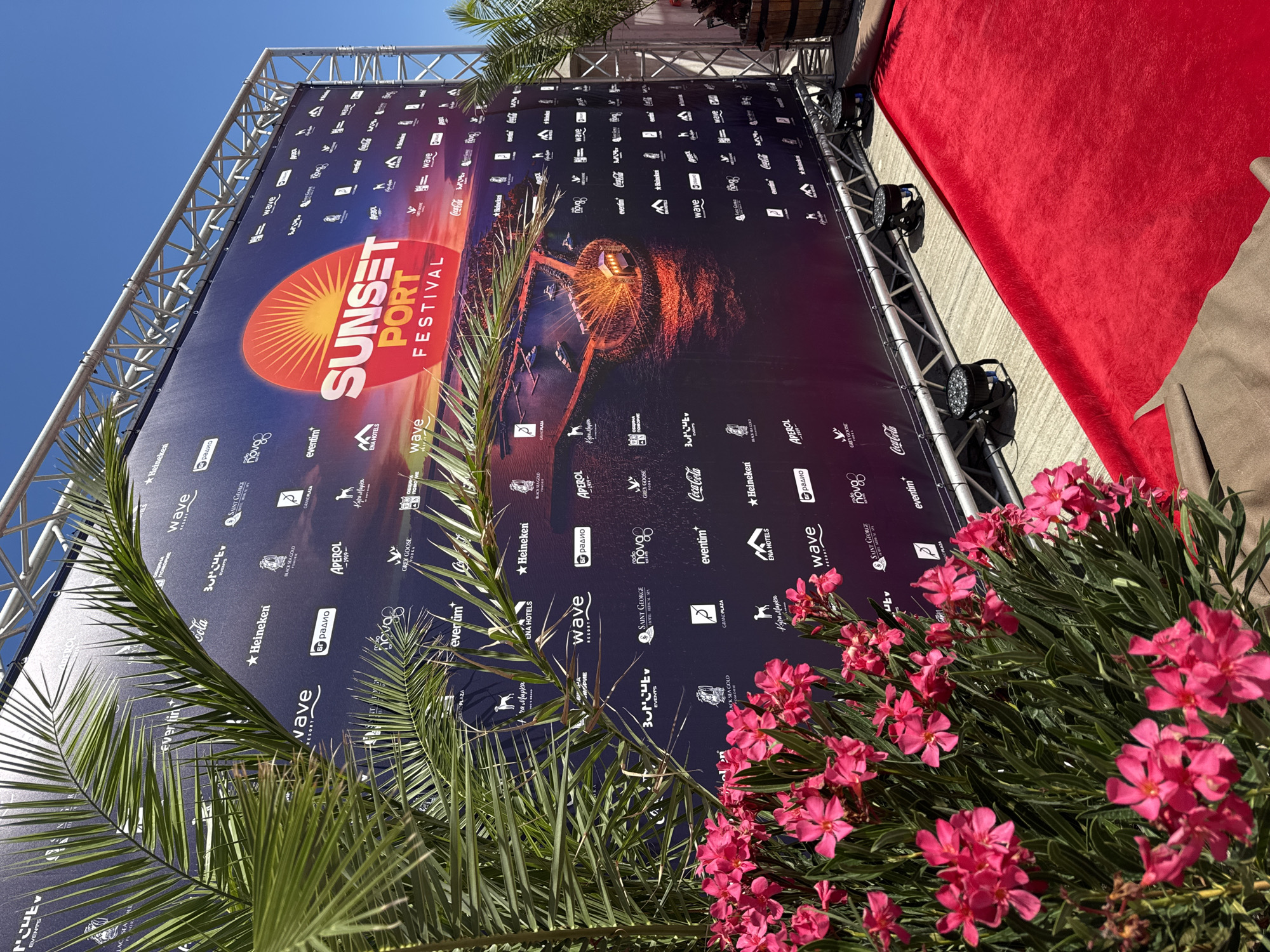 The image depicts a red carpet event setup in front of a large branded banner in Pomorie, Bulgaria. The banner advertises the "Sunset Port Festival" and features numerous sponsor logos such as Heineken, Coca-Cola, and Aperol. Palm fronds and vibrant pink oleander flowers frame the left side of the scene, creating a tropical ambiance. The banner is supported by a metal truss structure, and stage lights are visible on the ground. A wooden barrel is positioned on the far right, possibly for decoration or as part of the event setup. The ground is paved, and the overall scene suggests an outdoor festival or premiere event.