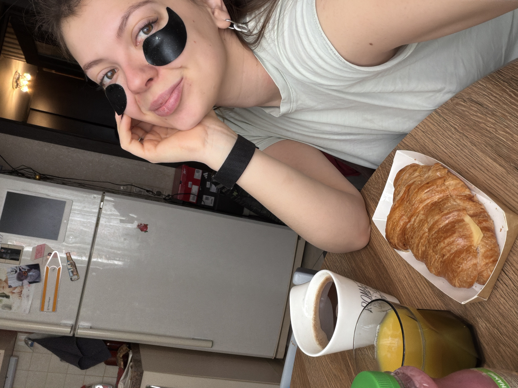 A young woman is captured in a close-up, angled selfie, appearing to be in a domestic setting in Khimki, Russia. She has brown hair, fair skin, and is wearing a light-colored, sleeveless top. She is engaged in a self-care routine, evident from the black, crescent-shaped under-eye patches applied below both her eyes. Her expression is calm and relaxed, with a subtle smile. She rests her chin on her right hand, which displays a ring on her ring finger and a black wristband or watch. A silver-colored earring is visible in her left ear.
The image suggests a morning scene, as indicated by the breakfast items laid out on a wooden table. To her right, there's a golden-brown croissant, presented in a paper tray, seemingly with a slice of cheese or butter tucked inside. Next to it is a white mug containing a dark beverage, likely coffee, and a clear glass filled with orange juice. A pink bottle, partially visible, has the text "HAMMARB" or "HAMMARBY" printed on it.
In the background, a white two-door refrigerator is visible, adorned with various magnets, photos, and a tablet mounted on its surface. Notable items on the fridge include a yellow pencil-shaped object and a miniature beer bottle magnet. A warm-toned light fixture can be seen in the upper left background, suggesting indoor ambient lighting. The overall atmosphere is casual and relaxed, indicative of a quiet breakfast and self-care moment at home.