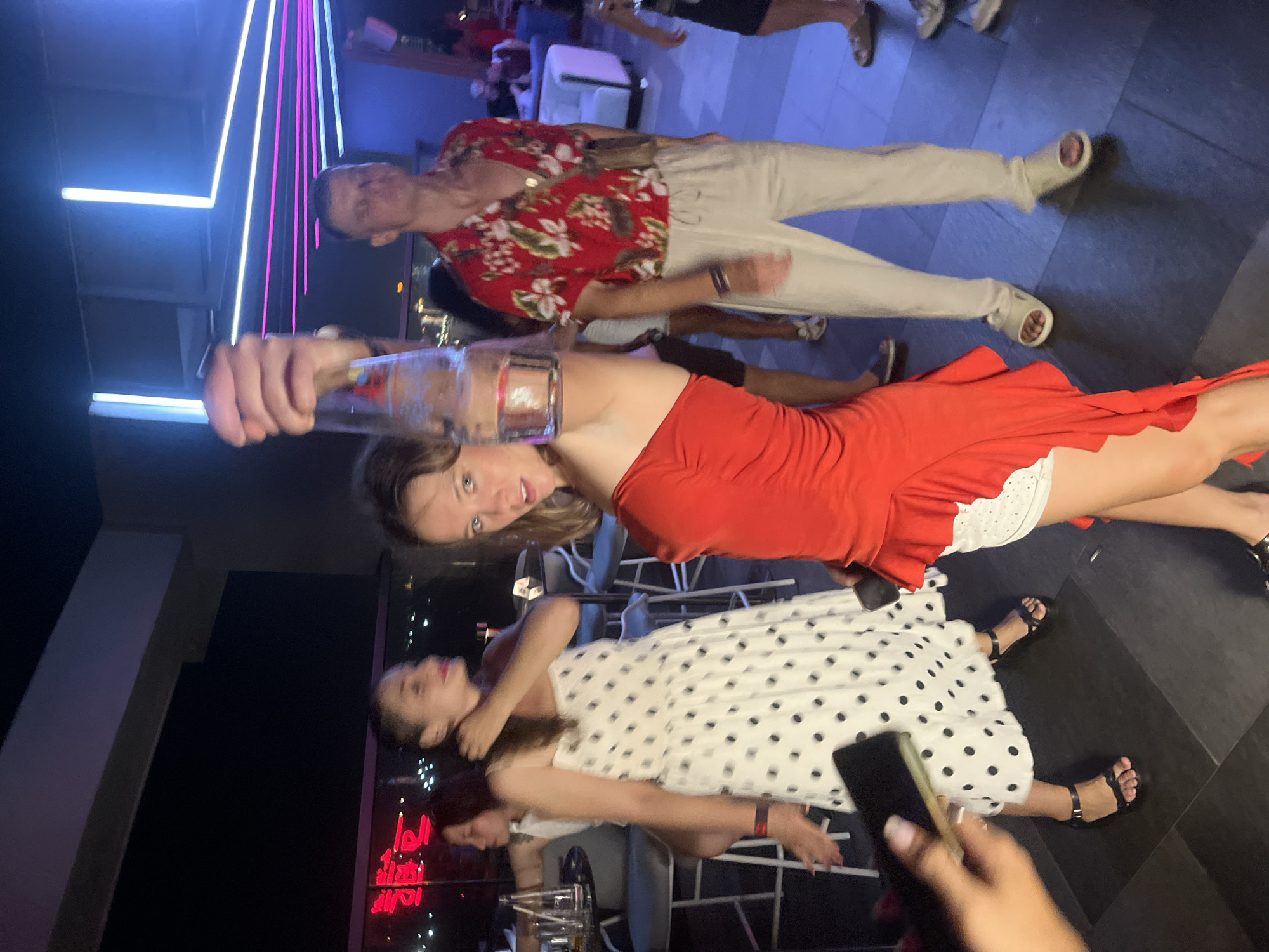 This image captures a group of people at what appears to be a lively evening event, possibly a bar or club, in Đà Nẵng, Vietnam. The setting features a dark floor with geometric patterns, illuminated by neon lights in blue and pink hues, suggesting a modern and energetic atmosphere.
The central focus is on three women. The woman in the foreground, wearing a red one-shoulder dress with a white lace hem, is holding up a glass of beverage. Her expression is engaging as she looks towards the camera. Next to her, another woman in a white dress with black polka dots is positioned lower in the frame, also looking towards the camera with a slight smile. To the left, a third woman, with darker hair, is partially visible.
In the background, a man wearing a vibrant red and white floral shirt and light-colored pants is also visible, looking towards the camera. Other individuals and elements of the venue, such as seating and lighting fixtures, are subtly present, adding to the sense of a social gathering. The overall mood seems celebratory and relaxed, with individuals enjoying themselves. The time of day is clearly night, indicated by the artificial lighting and the absence of natural daylight. No specific weather conditions are apparent. Some red neon lettering is visible in the lower left portion of the image, but the text is not legible.
