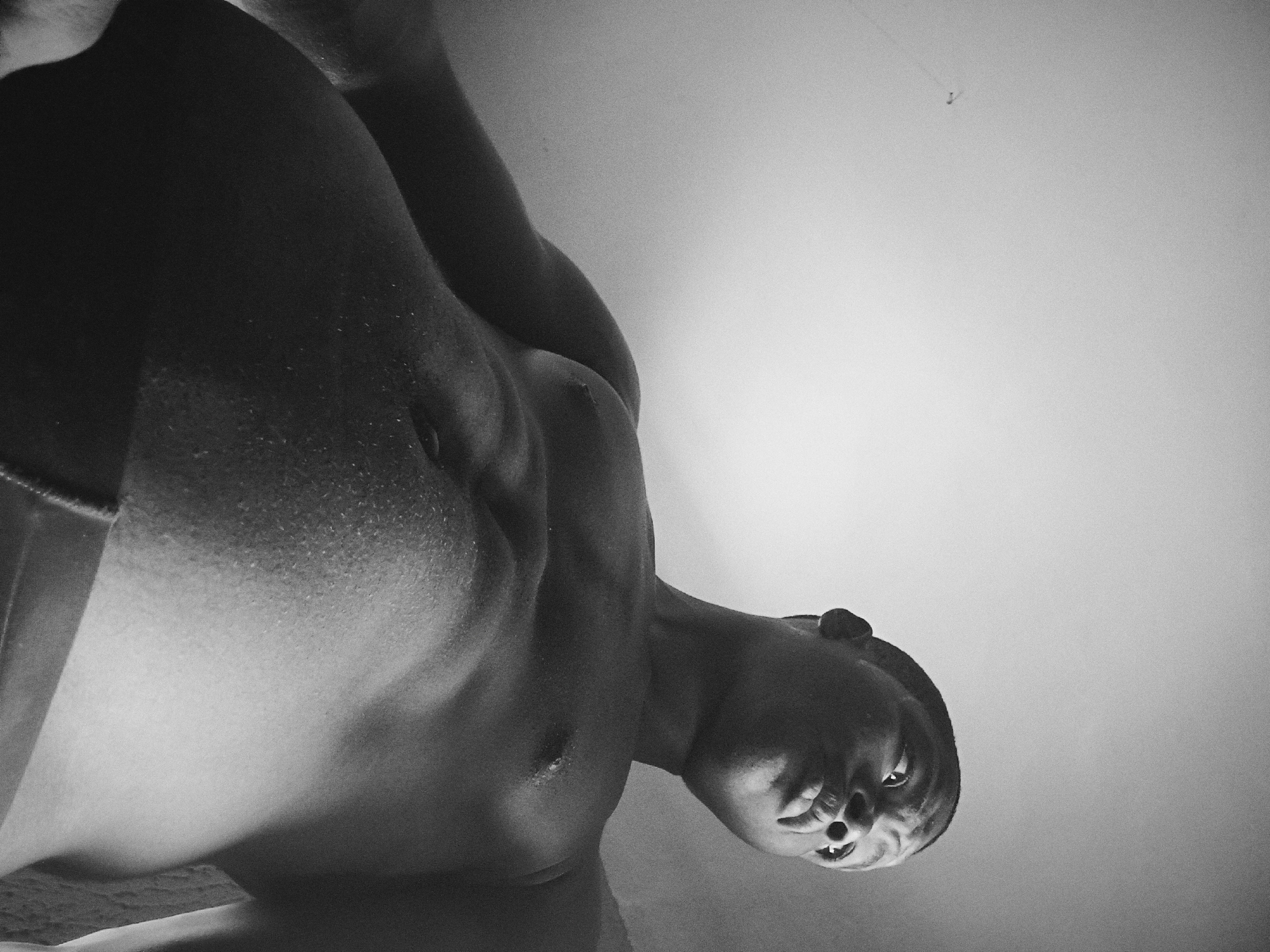 A black and white, low-angle photograph depicts a shirtless person, presumably male, with their body positioned closer to the camera and their head angled down towards the viewer. The person's face is visible, and their mouth is slightly open, revealing teeth. The person appears to have a dark complexion. The torso is visible with some sweat and the contours of the chest and stomach. The background is a plain, light-colored surface, likely a wall. A small hook is visible in the upper right of the frame. No specific clues indicate the location is Nkpor, Nigeria.
