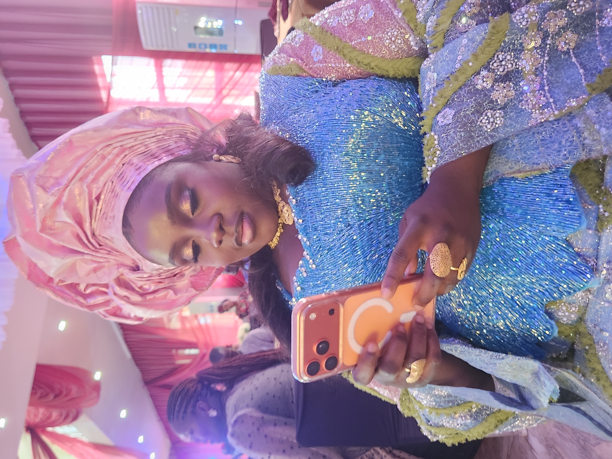 In Barden Yana, Nigeria, a woman is depicted holding and looking at a smartphone. She is wearing a vibrant, heavily embellished blue dress with iridescent sequins and a matching pink headscarf. The dress features intricate beadwork and patterned sleeves. The woman is adorned with gold jewelry, including a necklace and two statement rings. The background suggests an indoor event with draped pink fabric and soft lighting.