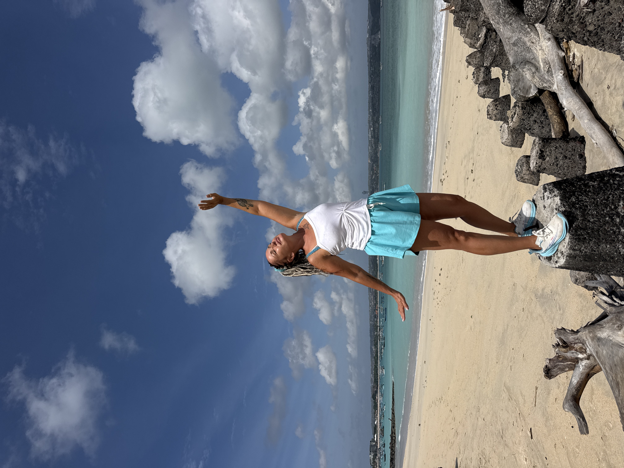 A woman with blonde braided hair and a white tank top and turquoise shorts stands on a rocky outcropping on a sandy beach. She is reaching her right arm up towards the sky, with her head tilted back and eyes closed. The ocean is a turquoise color and stretches out to the horizon where boats are visible. The sky is a bright blue with fluffy white clouds. In the foreground, driftwood is scattered on the sand, with some pieces forming a rocky barrier. The scene appears to be a tropical beach location.
