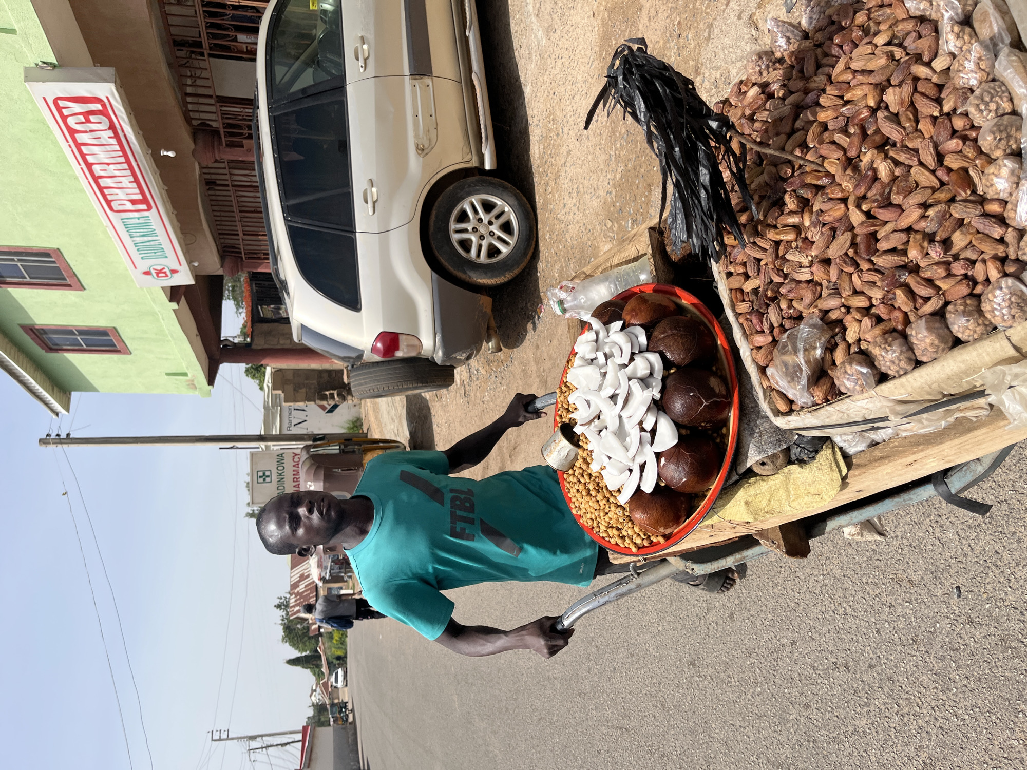 A medium shot depicts a man standing next to a cart of food for sale, on a paved street in Jos, Nigeria. The man is dark-skinned, with a shaved head, and is wearing a light green t-shirt. He is holding the cart's handlebars. The cart contains a red bowl with coconuts, chickpeas, and sliced coconut meat. Next to the bowl is a pile of dates and packages of peanuts. Behind the cart is a white SUV parked on the side of the street. Across the street is a pharmacy with a sign that reads "DADINKOWA PHARMACY." The background includes buildings, cars, and power lines under a bright sky. The image is rotated 90 degrees counterclockwise.