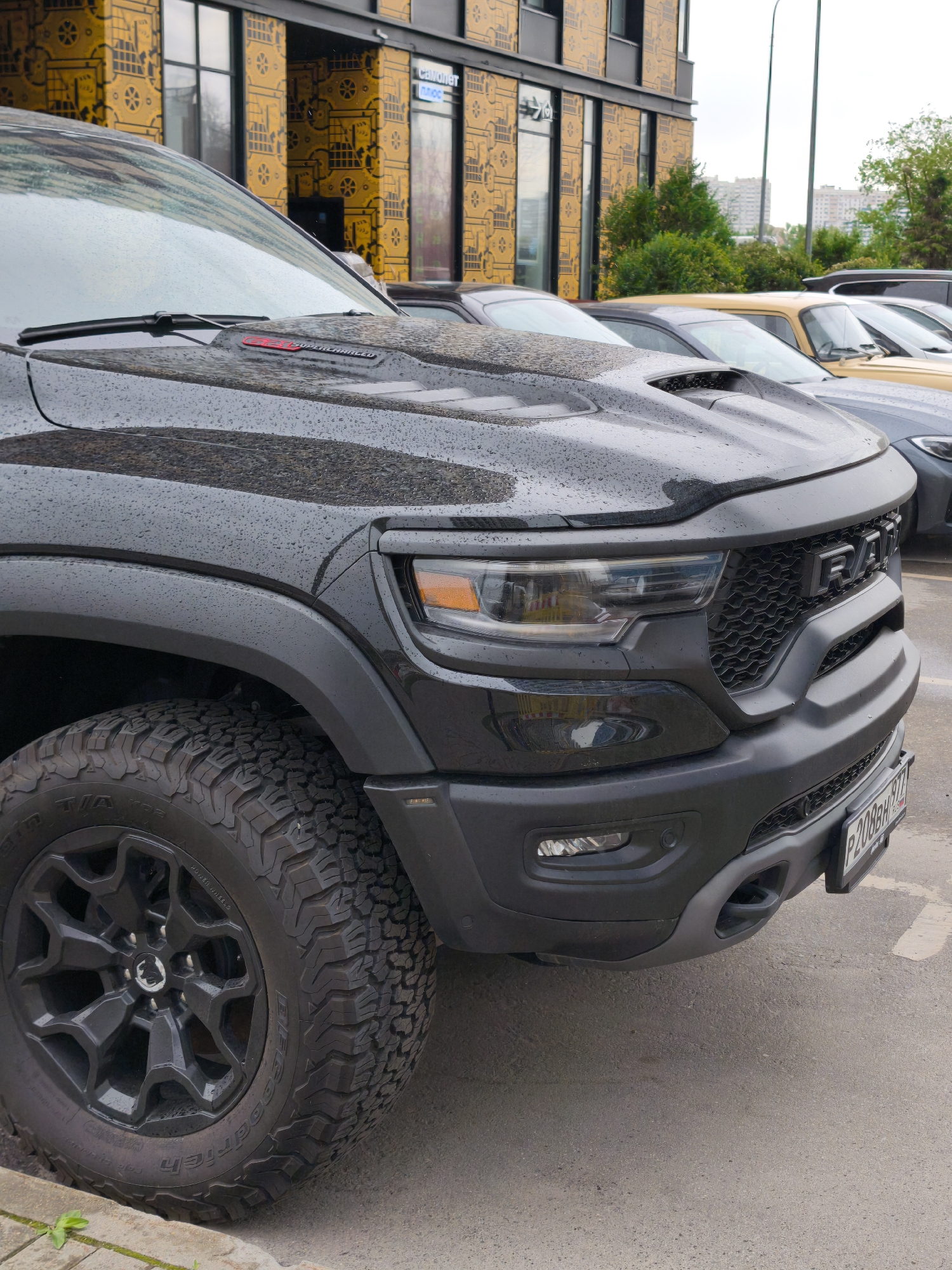 The image shows a black Ram TRX pickup truck parked in front of a building in Moscow, Russia. The building has a distinctive yellow and black patterned facade. Other parked vehicles are visible in the background, including a gray BMW and a yellow Lada. The ground is paved and appears wet, with raindrops on the truck. The license plate on the Ram TRX is a Russian plate.