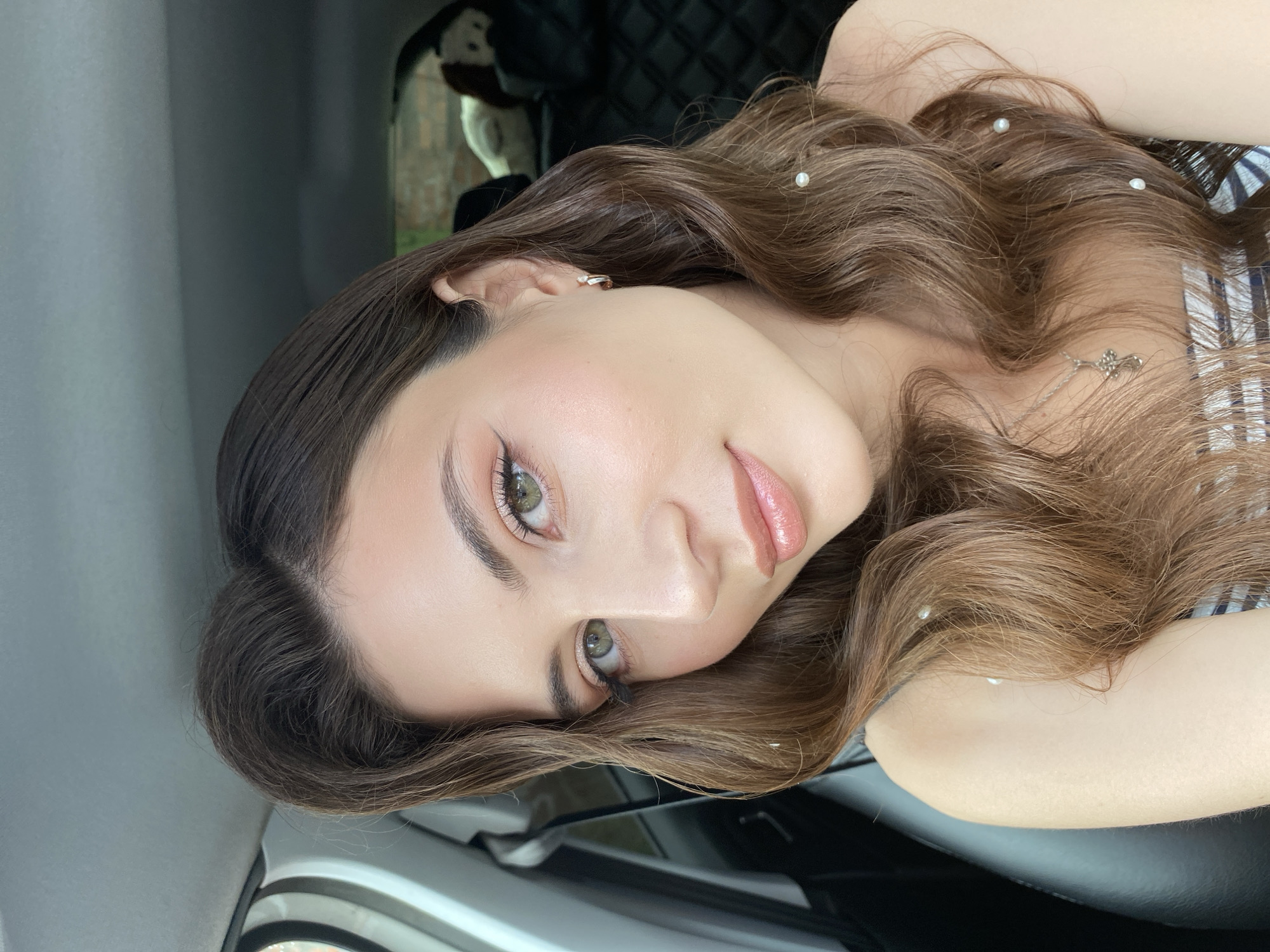 A young woman with long, wavy brown hair and green eyes is shown in a close-up selfie taken inside a car. She is wearing makeup, including eyeliner and lip gloss, and a silver necklace. Her hair is adorned with small white pearls. The background of the car interior is visible, including the car's ceiling, seat, and the window. The image does not contain any information that could place it in Votkinsk, Russia.