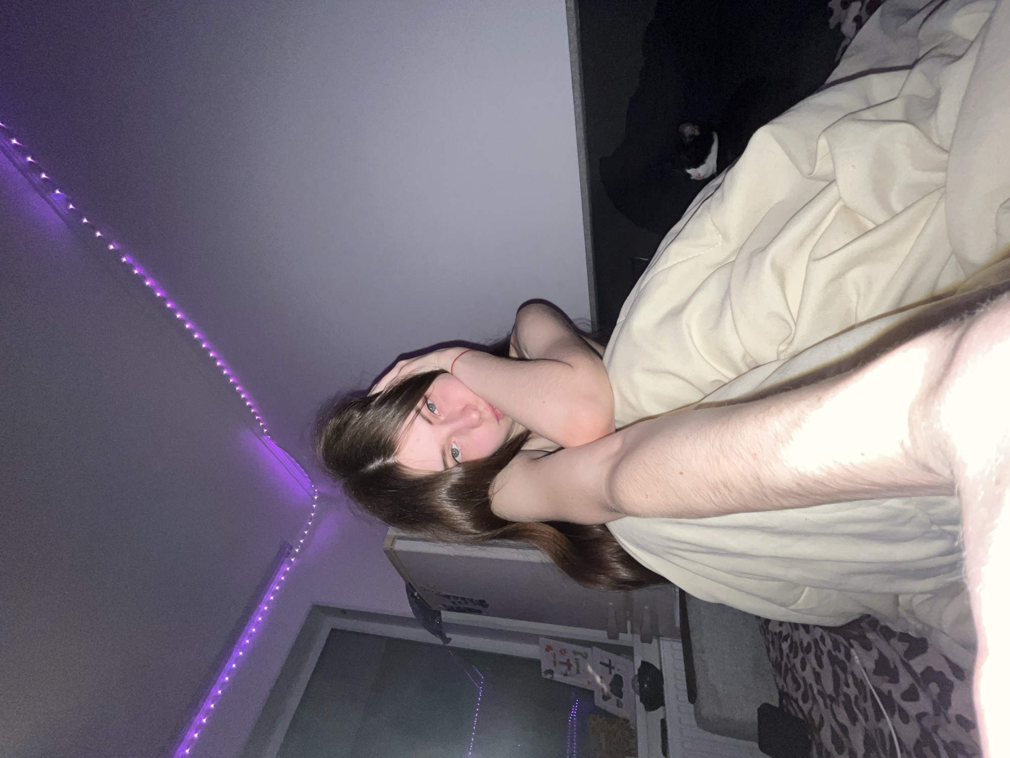 A young person is lying in bed under a beige blanket with dark hair across their face and green eyes open. Their arm is raised to their head with a red bracelet on their wrist. In the upper right corner, a black cat is partially visible. A string of purple LED lights runs along the wall on the left side of the frame. In the background, a window and items on a shelf are visible. The location is unknown and cannot be determined to be Saint Petersburg, Russia.