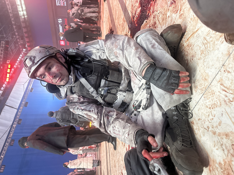 An indoor film set in Huangdao District, China, depicts the aftermath of a violent event. A Caucasian male, appearing to be in his twenties, is seated cross-legged on a simulated ground textured with artificial bloodstains and pools. He is dressed in a light grey camouflage tactical uniform, a grey tactical vest, a grey tactical helmet, and black fingerless gloves, all heavily splattered with artificial blood across his face, helmet, uniform, and hands. A black headset is visible around his neck. He gazes upwards and to his left. Between his legs rests a white object with red and green markings. In the background, a large blue screen occupies the left side of the set. Several individuals are present: a man in a dark, blood-stained suit walks away from the foreground subject towards the blue screen, while other figures, including one in a "SECURITY" labeled vest and a woman in a white coat, are visible. On the right, a group of people, some in tactical gear and others in civilian attire, are gathered near a large red display screen bearing the text "203" and "SUPPLY AREA". Overhead, metal rigging supports arrays of bright red stage lights, casting a strong red illumination across the scene, particularly on the right side.